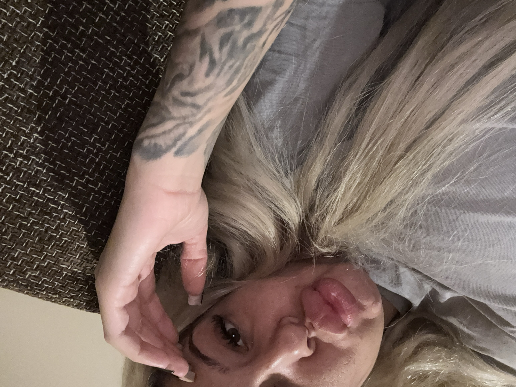 A close-up, downward-facing shot depicts a woman lying down. Her blonde hair is spread around her head. She has one arm raised, with a tattoo visible on her forearm. Her lips are pursed, and her tongue is slightly visible. A brown patterned textile is in the background. The location is Unknown, Unknown.