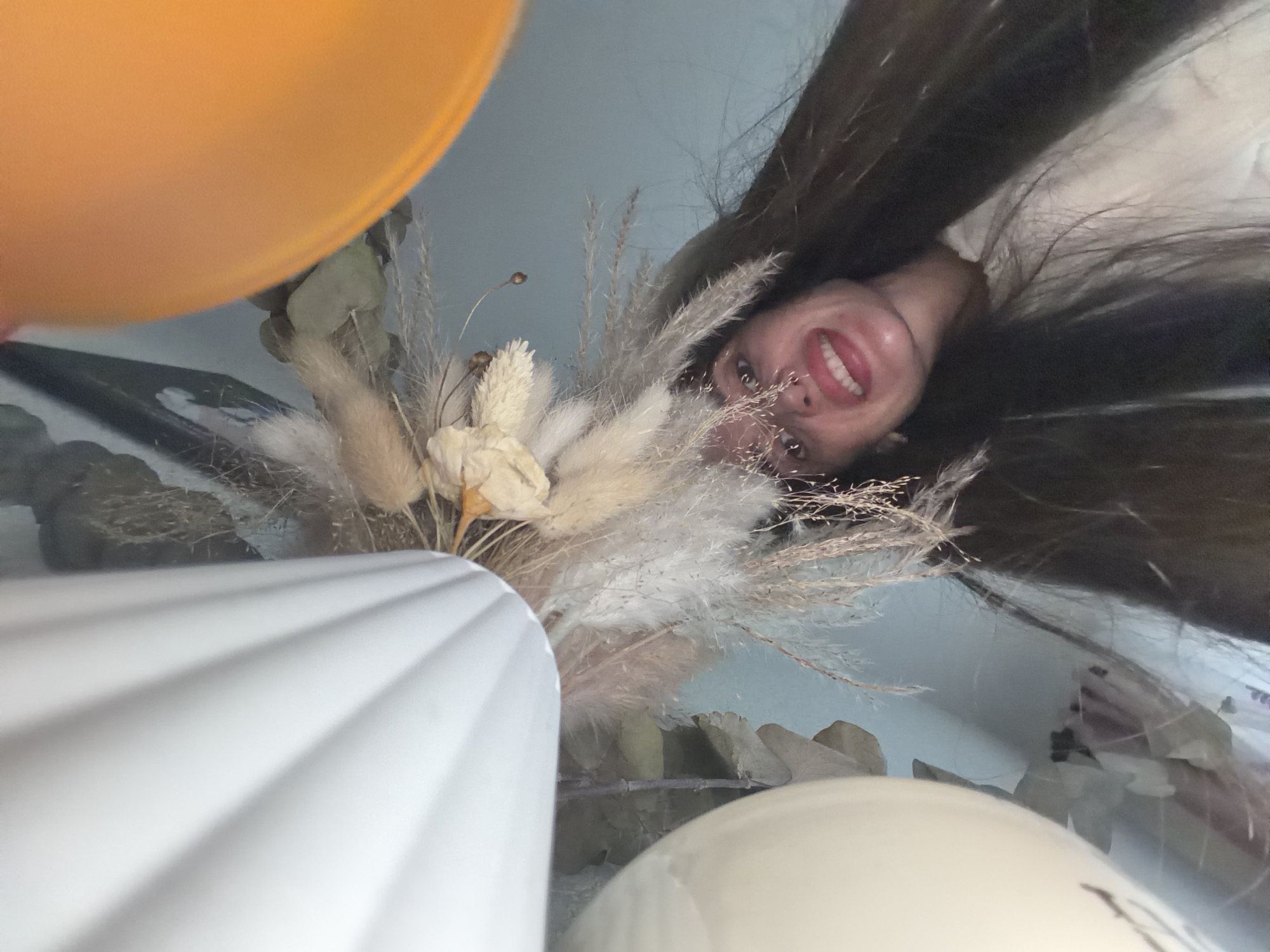 A low-angle shot captures a woman smiling toward the camera. She has long, dark hair. Framing her face is an arrangement of dried flowers in a tall, white vase, partially obscured in the foreground. An orange object is visible in the upper left, with a tan-colored spherical object in the lower center. The backdrop is a light-colored wall. The location in the city of Saint Petersburg, Russia, is not discernible.