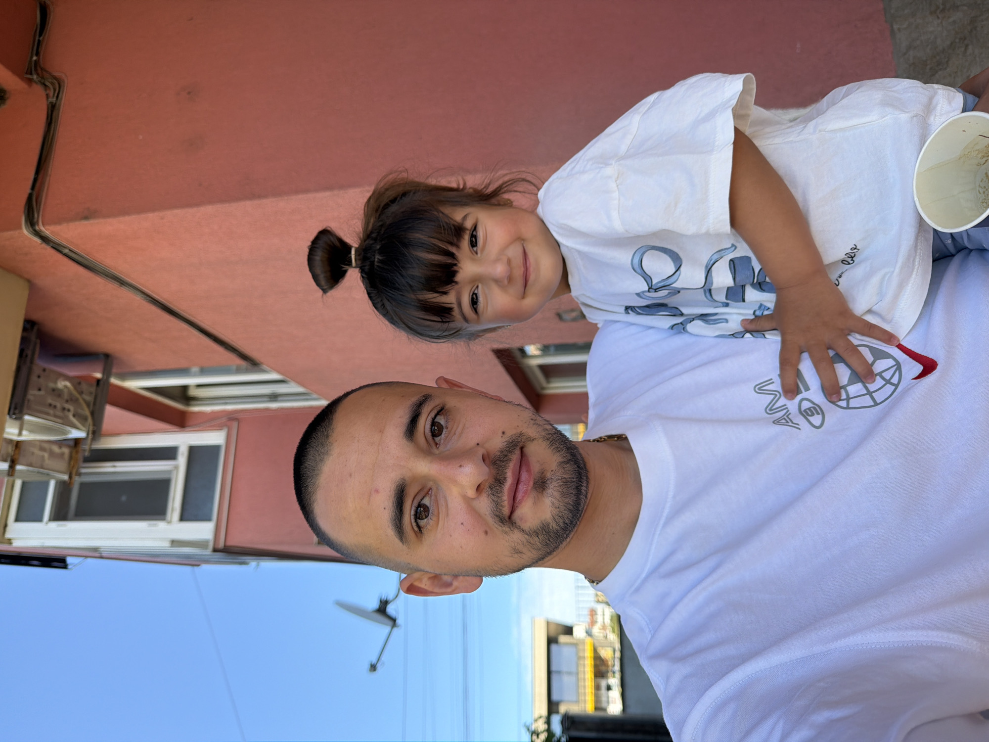 A father and daughter are posing for a selfie outdoors in front of a coral-colored building. The father is on the left, his face close to the camera, with a shaved head and a short beard. He is wearing a white t-shirt with a graphic design on it, and a gold chain. The daughter is on the right, being held by the father. She has her hair pulled up in a small bun and is wearing a white t-shirt with blue ribbon-like graphics. She is smiling at the camera. The background shows the coral building, a clear blue sky, and a satellite dish mounted on a tower. The overall lighting suggests it is daytime, likely with bright sunlight. The location is specified as Burgas, Bulgaria, though this information is not visually discernible from the image itself.