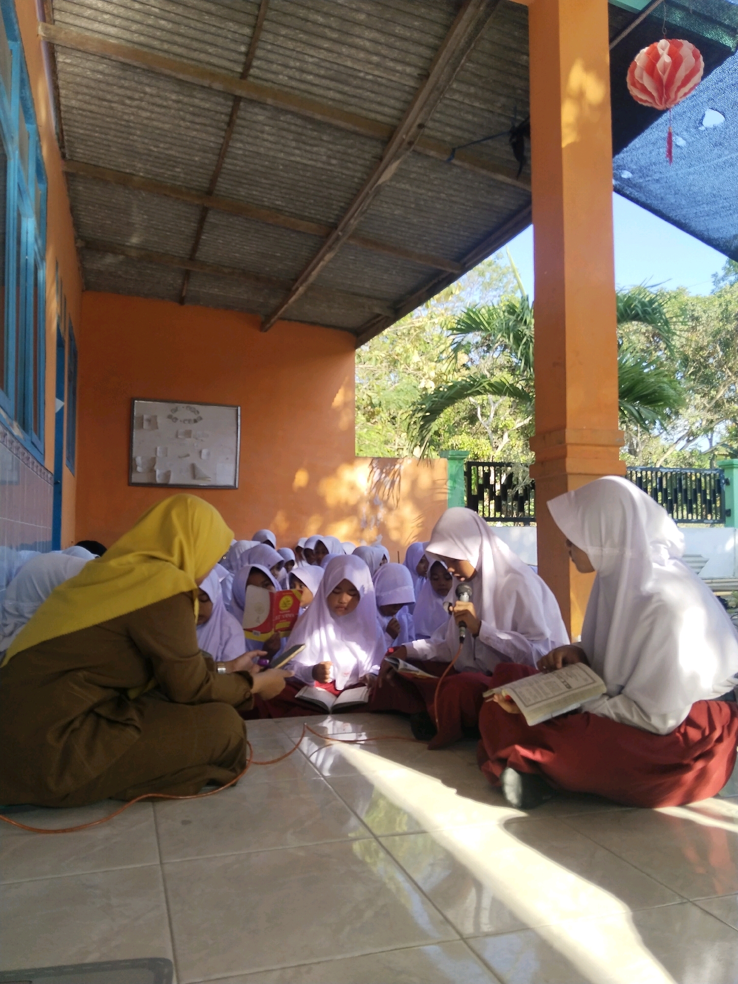 A group of young Indonesian students, mostly female, are gathered outdoors, sitting on a tiled floor. They appear to be in a classroom or school setting, possibly in a covered walkway or veranda, as indicated by the corrugated metal roof overhead and an orange-painted wall. Many of the students are wearing white hijabs and uniforms, suggesting a religious school or a lesson with a religious component. Some are reading books, while others seem to be listening. One student is holding a microphone, perhaps reciting or singing. A teacher, wearing a yellow hijab and a brown tunic, is seated to the left, looking at a mobile phone and holding a cord that runs to the microphone. In the background, there are trees, a fence, and a bright blue sky. A decorative red and white lantern hangs from the roof on the right side of the image. The scene is illuminated by sunlight, creating strong shadows on the floor. The overall impression is one of a learning environment, likely a school in Taraban, Indonesia.