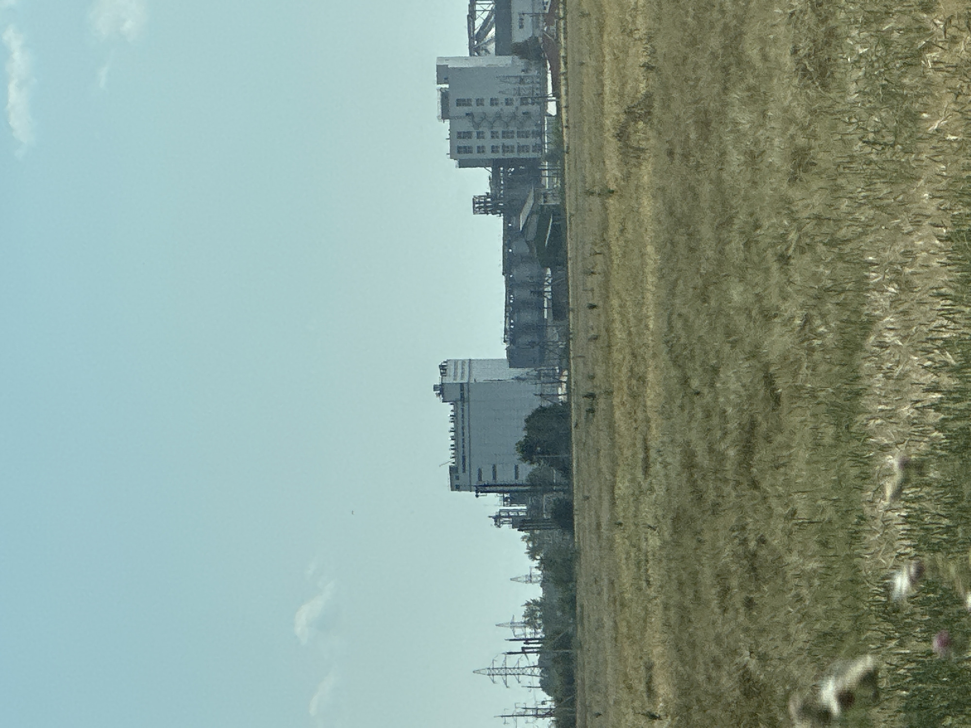 An eye-level, medium shot captures a vast, dry field of golden-brown grass in the foreground under a clear, pale blue sky. In the middle ground, a sprawling industrial complex dominates the landscape, featuring multiple large, grey, rectangular buildings, some with visible windows and structural details. Several tall, lattice-like electrical pylons stand to the left of the industrial buildings. The background is a hazy, pale blue sky with a few small, wispy clouds. The scene appears to be set in an industrial zone on the outskirts of a city.