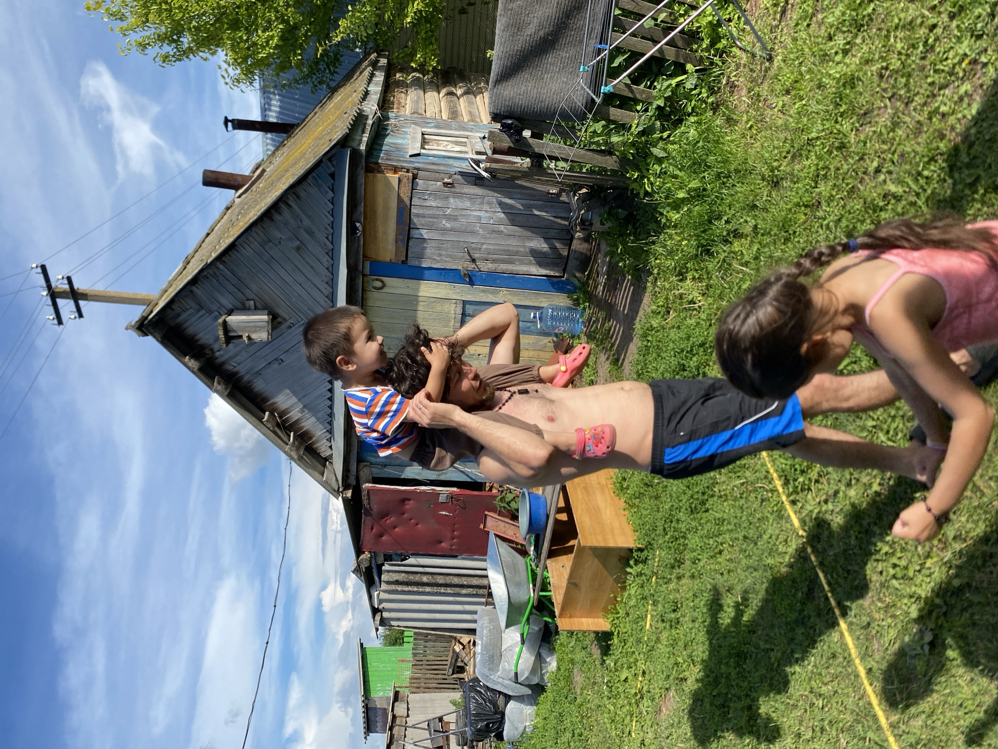 In the village of Chirsha-Tartysh, Russia, a shirtless man with a beard carries a young boy on his shoulders. The boy wears a striped t-shirt and pink Crocs. A younger girl, with dark hair in a braid and wearing a pink tank top, is in the foreground, her back to the camera. Behind them stands a weathered wooden house with a metal roof and a red door. Various objects are scattered around the yard, including a wheelbarrow, a wooden box, and a clothes drying rack. The ground is covered in green grass, and the sky is blue with scattered white clouds.