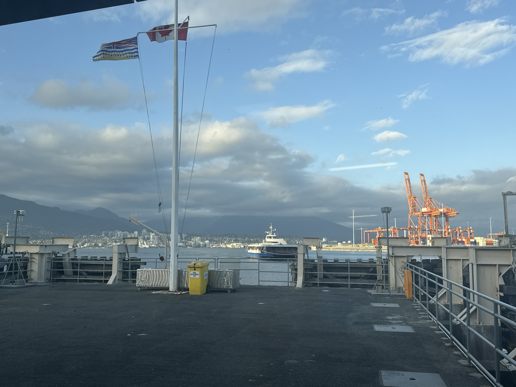 The image depicts a pier or ferry terminal in Vancouver, Canada, identifiable by the presence of the British Columbia flag and the Canadian flag on a flagpole. In the background, a cityscape of Vancouver is visible, framed by mountains. A vessel transits on the water. The foreground is composed of the pier surface, railings, and maritime structures, including a yellow "Spill Response Kit." Container cranes are visible in the middle distance. The sky is partly cloudy. No people are visible.