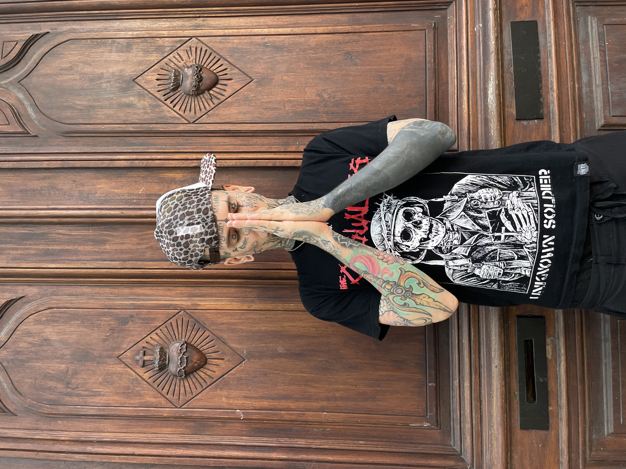 A person with extensive tattoos on their face, neck, and arms is standing in front of a large wooden door. The person is wearing a black t-shirt with a graphic print and a patterned cap. Their hands are pressed together in front of their face, in a prayer-like gesture. The wooden door features ornate carvings, including a heart with rays emanating from it. The setting appears to be outdoors, with natural light illuminating the scene. The location is in the city of Buenos Aires, Argentina.
