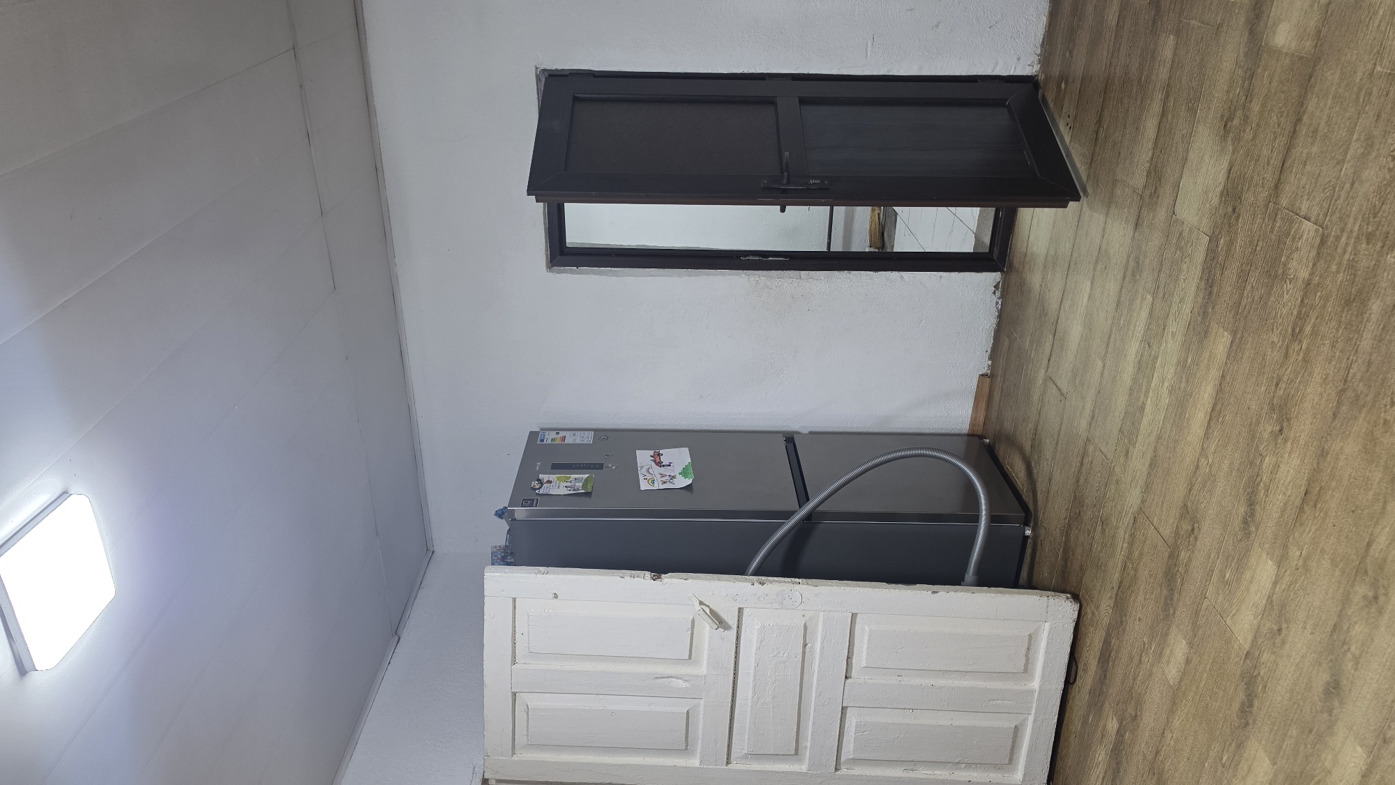 The image depicts an indoor scene, likely a kitchen or utility room, in Kochkor, Kyrgyzstan. The room features a light-colored ceiling with a modern, rectangular LED light fixture. The walls are painted white and appear to have a textured finish. On the left side of the frame, a tall, narrow, white wooden door is propped open, revealing its paneling. To the right of the door stands a modern, silver refrigerator, adorned with a few pieces of paper, possibly children's drawings. A flexible grey hose is attached to the base of the refrigerator and extends towards the floor. In the background, to the right, a dark brown door with a metal handle is partially ajar, leading to another room or area. The floor is covered with light brown wood-look laminate flooring. The overall impression is of a functional, somewhat sparsely decorated space.