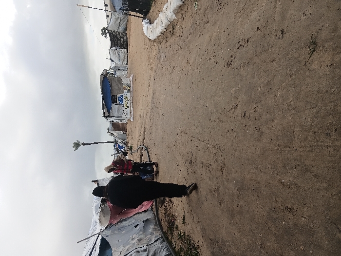 The image depicts a makeshift encampment in Khan Yunis, Palestinian Territory, under an overcast sky. The foreground features a dirt and sand path, showing minor undulations and sparse dry vegetation. A person wearing dark clothing, including a dark beanie or hood and a dark jacket, walks away from the camera along this path, with their left foot lifted mid-stride. Further into the scene, a second person, dressed in a light-colored hooded top and a red vest, stands near a temporary shelter, seemingly attending to a dark, low-lying object or cart.
The environment is characterized by numerous temporary structures constructed from tarpaulins, fabric, and corrugated materials. These shelters are predominantly white and grey, with some featuring blue tarpaulins or red fabric accents. A dark, flexible pipe or hose runs along the ground next to the leftmost shelter. In the mid-ground, a tall, slender palm tree stands solitary against the sky. On the right side of the path, a low barrier made of white sandbags or similar material defines an edge. The overall scene indicates a temporary settlement with basic, improvised housing.