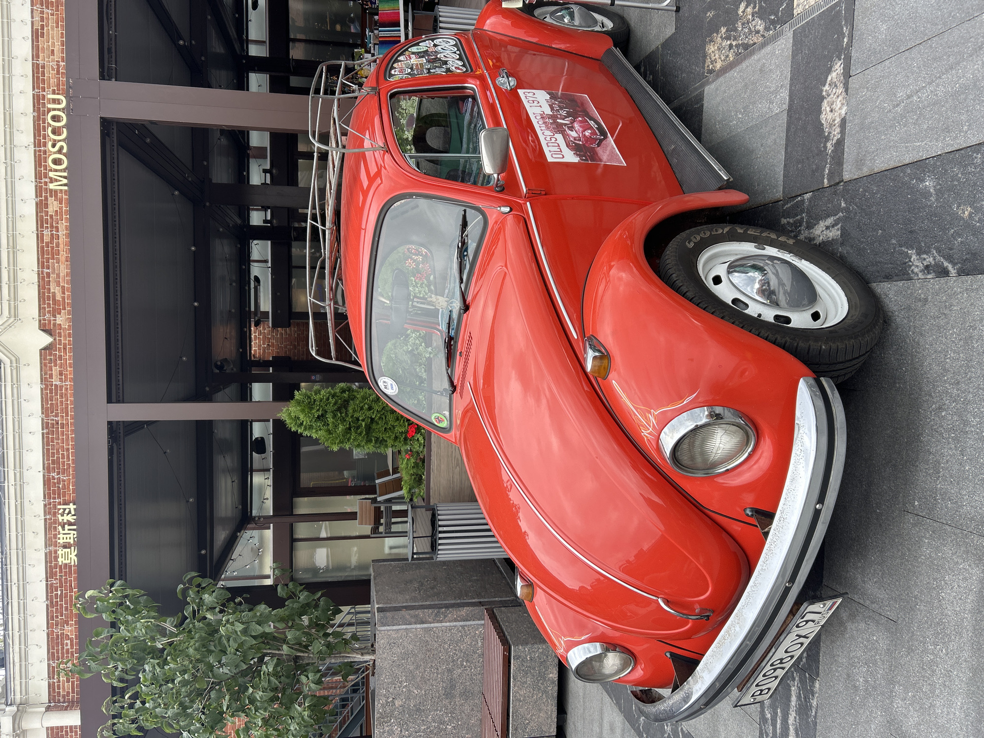 A red 1973 Volkswagen Beetle with a roof rack is parked on a grey stone-tiled pedestrian area in front of a building with red brick and brown trim. The building has a sign with Chinese characters and the word "MOSCOU" in English. The car has a license plate with the Russian abbreviation "RUS" and the characters "B098OX 97". There are stickers on the car's rear window and a larger sticker on the passenger door depicting an older version of the same car with the text "OLDSCHOOL 1973". A tree and some greenery are visible to the left of the car, and outdoor seating is visible through the building's windows in the background. The sky is overcast.