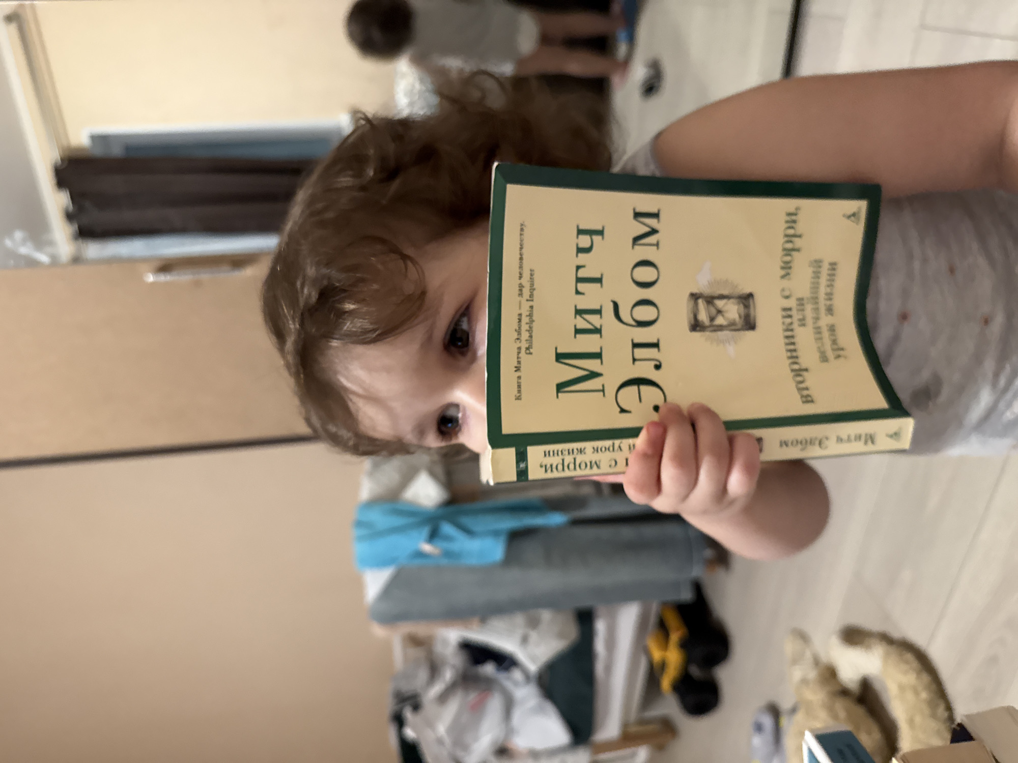 A young child with brown curly hair is holding a book titled "Mitch Albom" (in Russian) in front of their face, partially obscuring it. The book's cover is tan with green trim. The background shows a room with various objects, including bedding, clothes, and other household items. There are also glimpses of other people in the blurred background. There is no evidence in the image to suggest that the image was taken in the city of Tobolsk, Russia.