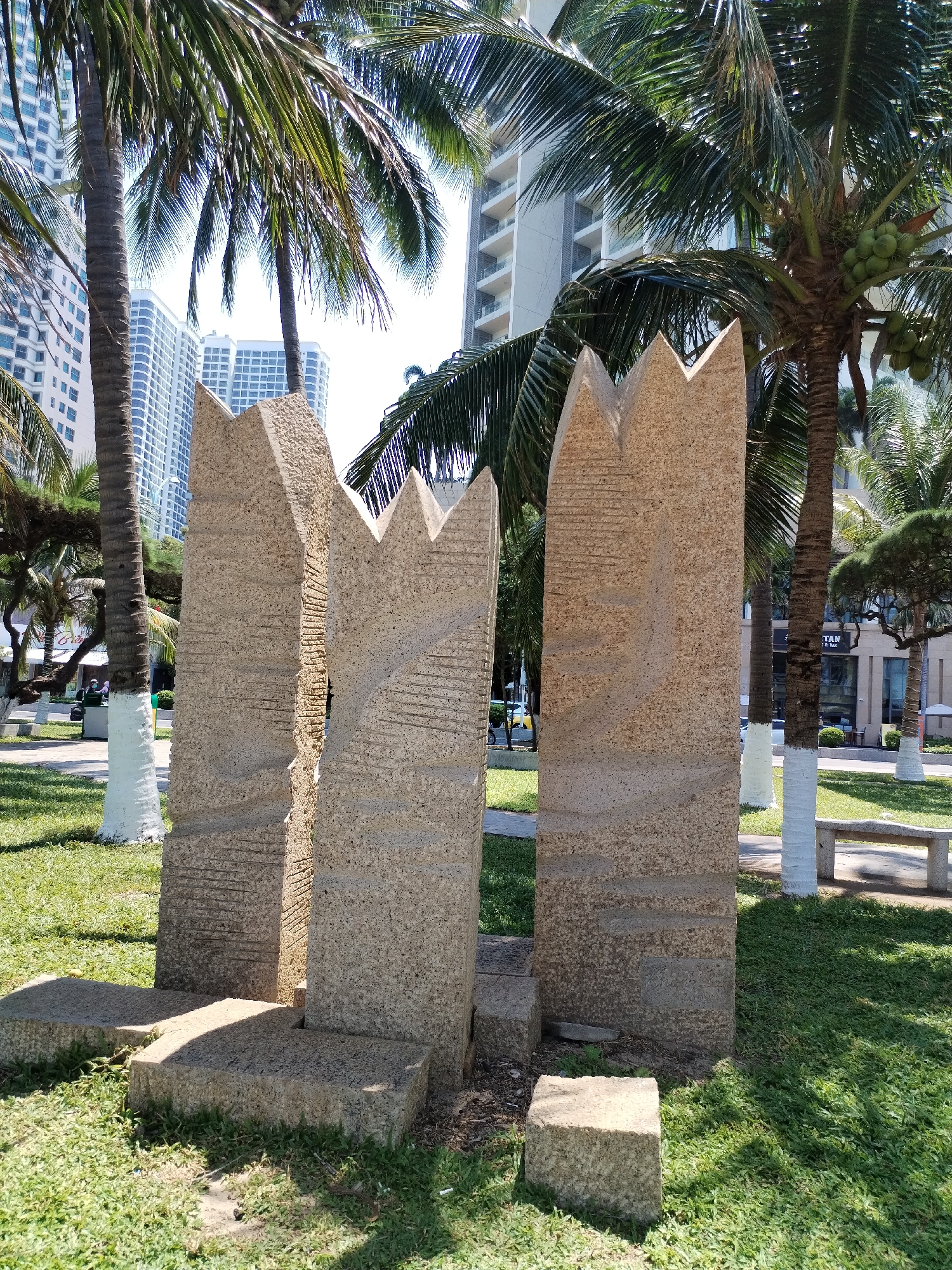 The photograph depicts a park in Nha Trang Ward, Vietnam. The scene features three roughly textured, rectangular stone sculptures with jagged, crown-like tops, placed on individual square stone bases. The sculptures are surrounded by a vibrant green lawn. Several palm trees with whitewashed trunks are scattered throughout the park, and tall buildings can be seen in the background. A stone bench is visible in the distance, adding to the park's scenery.