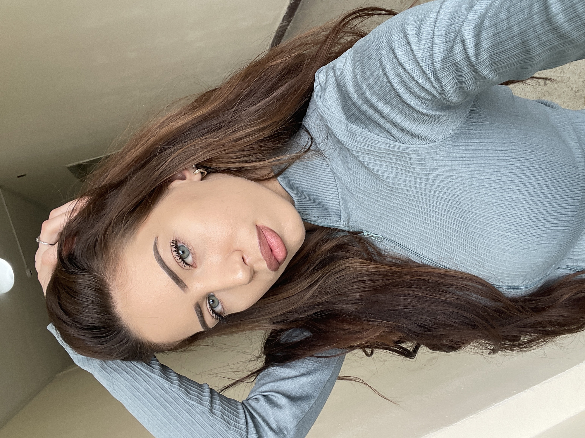 A young woman is depicted from a high-angle, close-up perspective. Her long, brown hair is styled loosely around her face and shoulders. She has fair skin, light-colored eyes, and wears neutral-toned lip color. Her expression is direct and engaging. She is wearing a light blue, textured long-sleeved shirt. The background appears to be a neutral-colored wall or floor. No specific context indicates the location being Veliko Tarnovo, Bulgaria. No other people, actions, or interactions are visible.
