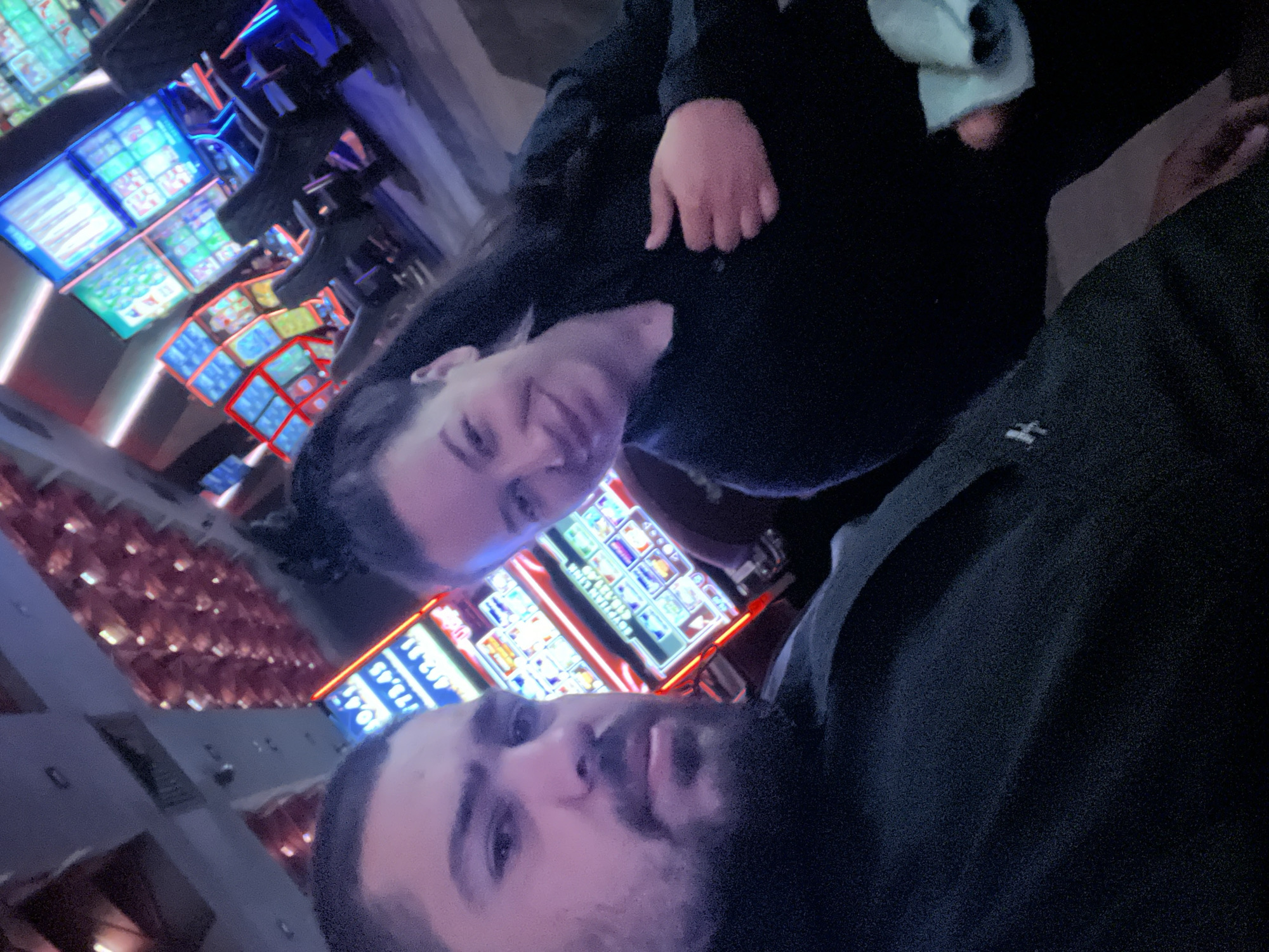 In a dimly lit indoor setting, a man with a beard and a woman with dark hair are taking a selfie. The background suggests they are in a casino, with rows of brightly lit slot machines visible. The slot machines have displays showing numbers and graphics. The woman is smiling, and the man is looking towards the camera. The woman is holding a light blue cloth in her right hand. The man is wearing a dark garment, and the woman is wearing a dark, long-sleeved shirt. The lighting is artificial and predominantly purple and blue, with red accents from the slot machines. The image does not provide any specific location within Burgas, Bulgaria.