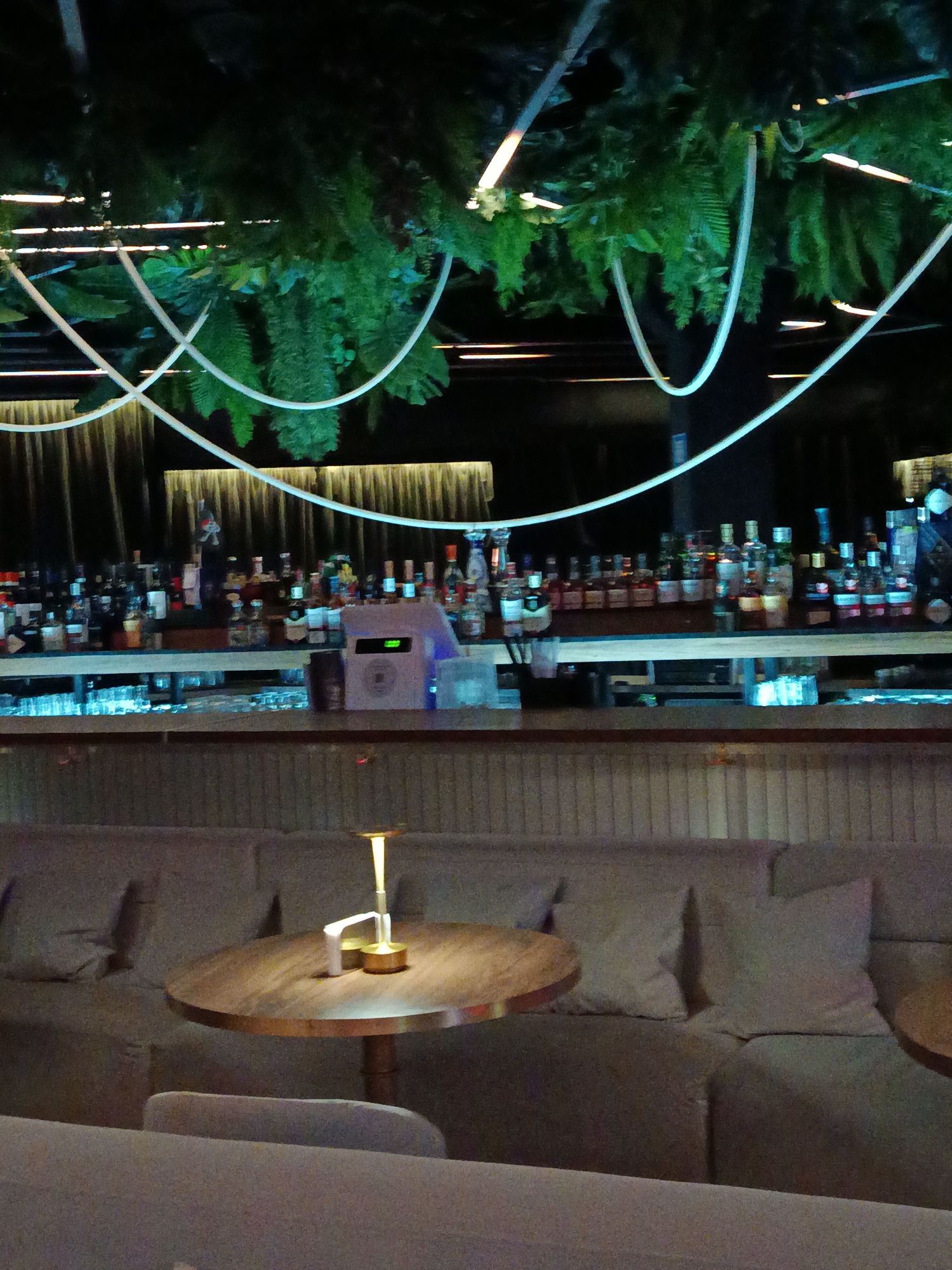 The image shows the interior of a dimly lit bar or lounge. Above the bar, there are decorative green plants and white curved light fixtures. Behind the bar, there are many bottles of liquor. In the foreground, there is a round table with a lit lamp on it, and a cushioned seating area. The exact location is not specified.