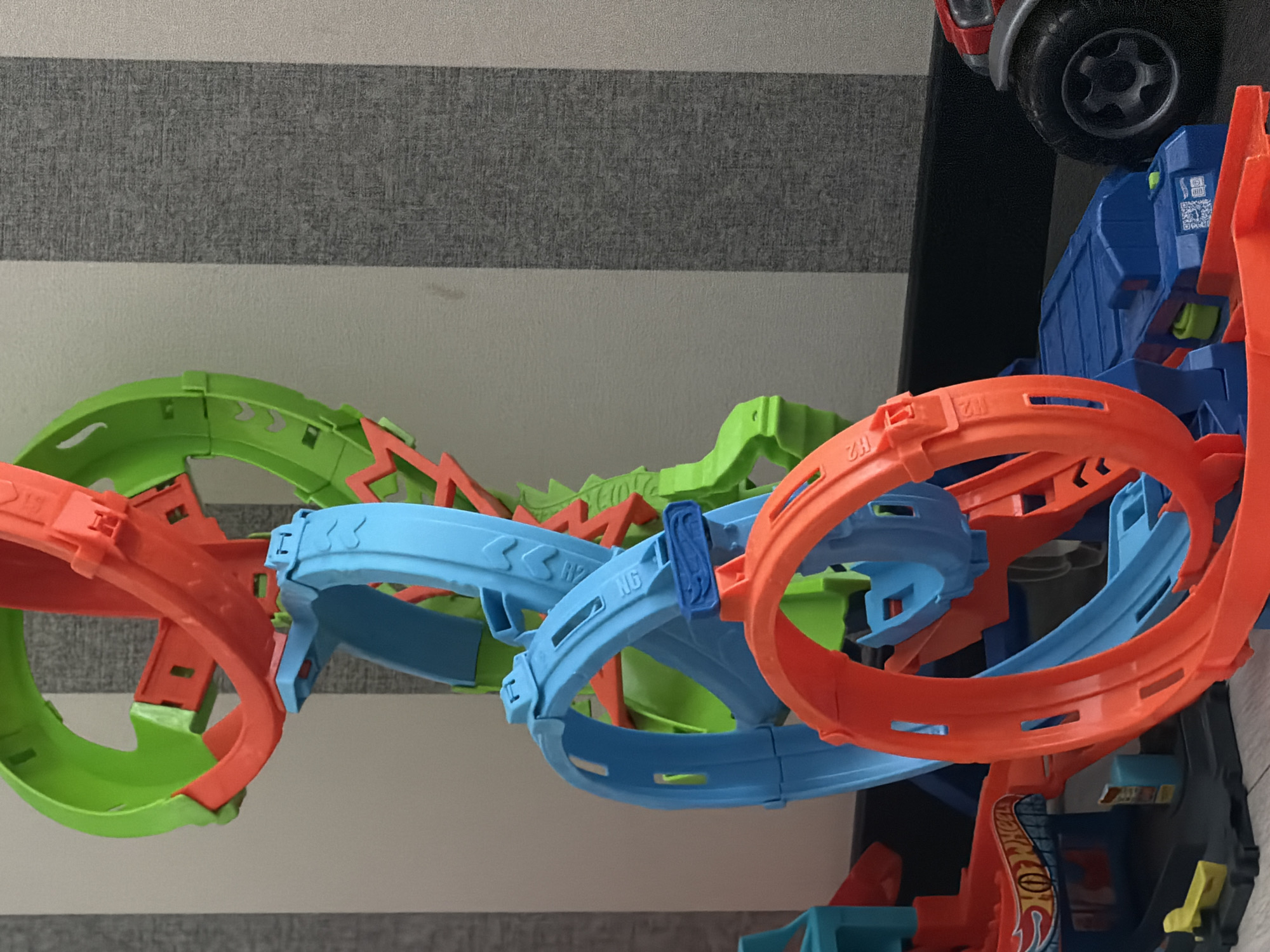 The image shows a close-up of a colorful toy car track set, predominantly featuring bright green, blue, and orange plastic pieces forming loops, curves, and ramps. The track is assembled vertically and is angled towards the right side of the frame. In the lower right corner, a portion of a toy vehicle with a large black tire and red body is visible. The background consists of a wall with alternating vertical stripes of light gray and a darker gray textured pattern. The lighting is soft and diffused, suggesting an indoor environment. The scene appears to be a still life of toys, with no discernible human presence or activity. The location of Bila Tserkva, Ukraine, is not visible or suggested by any elements within the image.