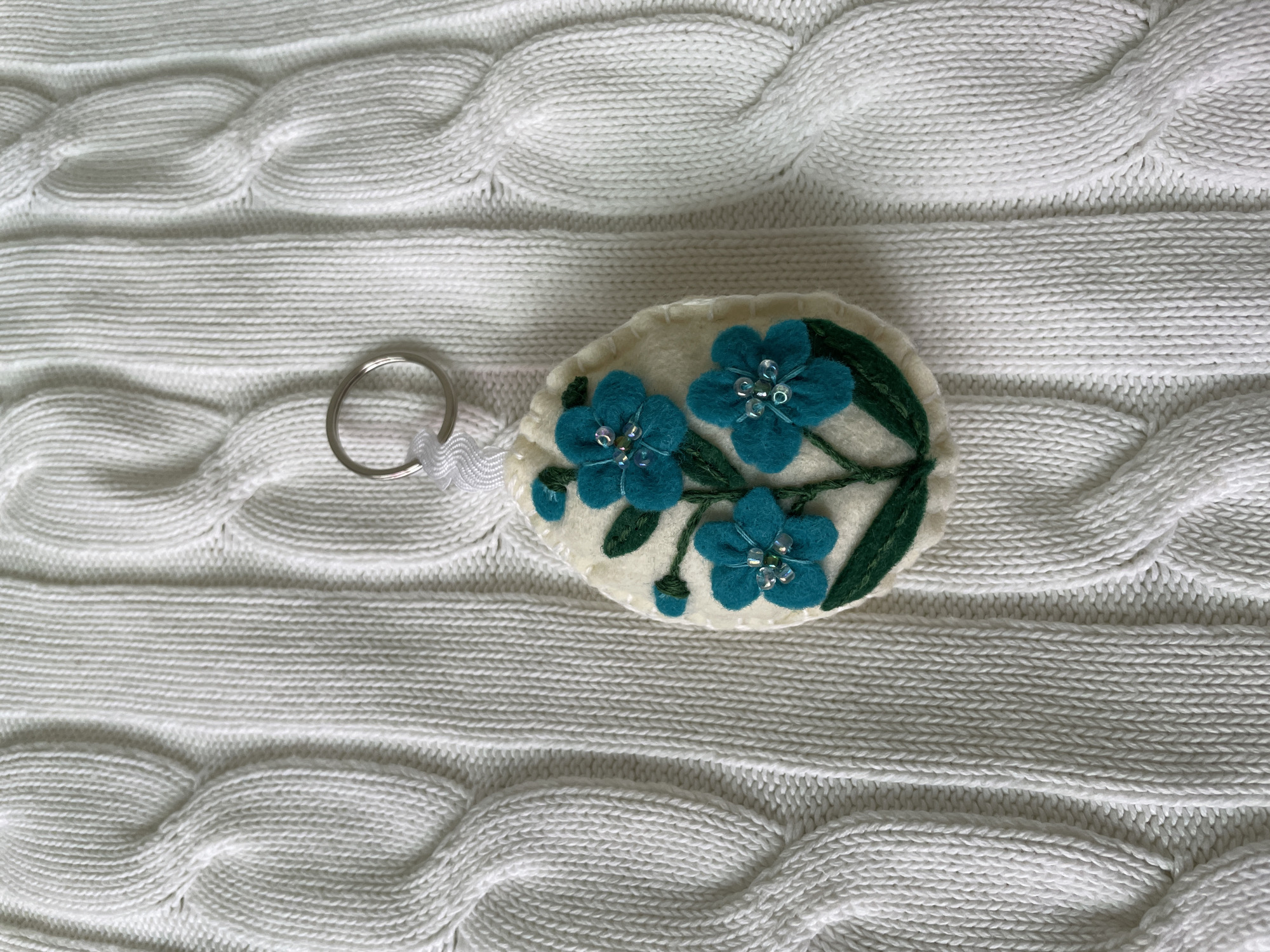 A handmade keychain with blue felt flowers is lying on a white cable knit sweater. The keychain is egg-shaped with an off-white felt background, and three blue flowers with beaded centers are sewn onto it. Green felt stems and leaves complete the floral design. A silver keyring is attached to the keychain with a short white ribbon. The sweater has a pronounced cable knit pattern with both horizontal and diagonal ridges. The photograph was taken in a place with an unknown street address in the city of Лесной Пейзаж Кп (шарапово, Russia.