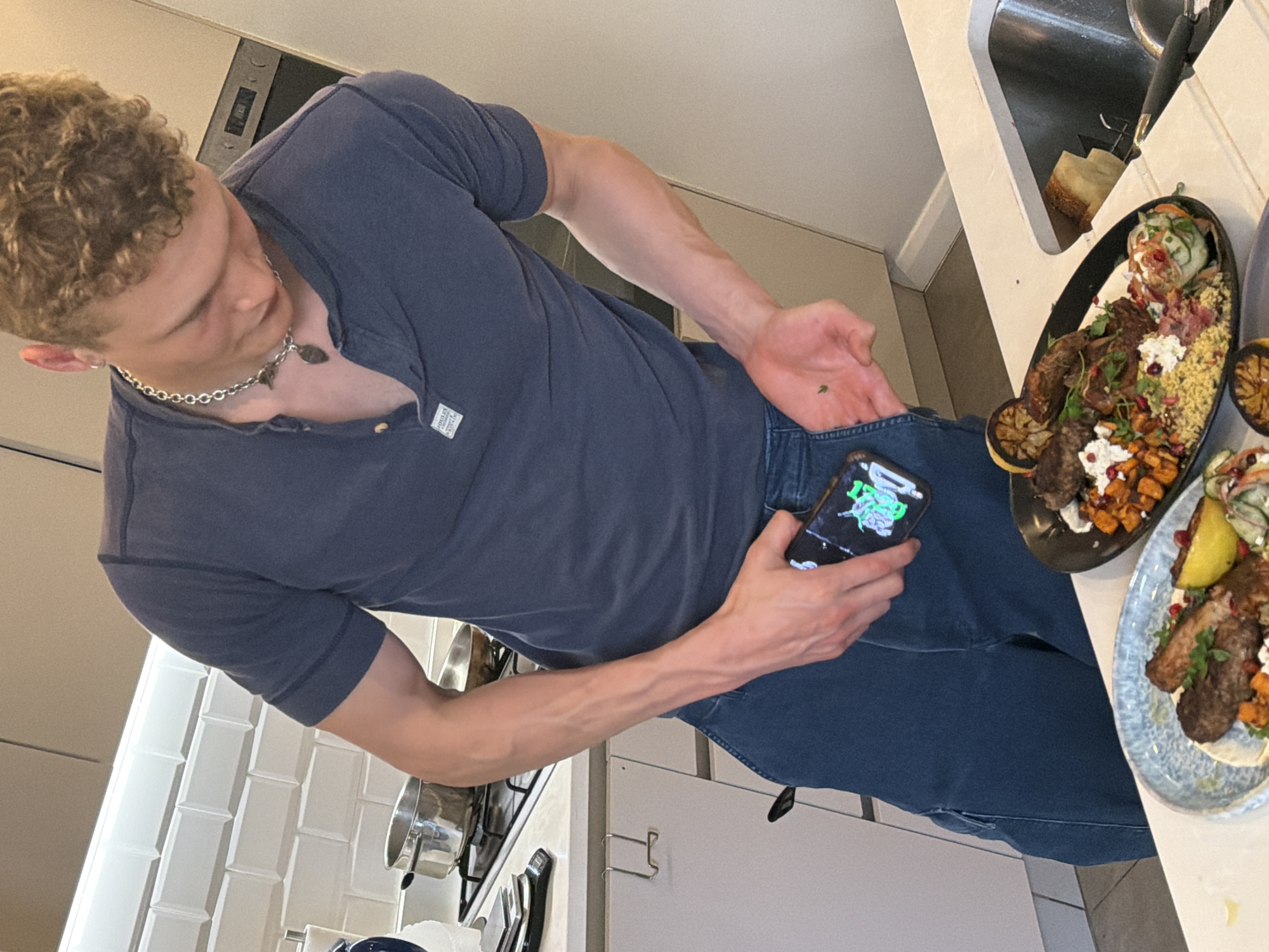 A man with curly blonde hair and a silver chain necklace is holding a cell phone in his left hand while standing in a kitchen. He is wearing a blue short-sleeved shirt and blue jeans. His right hand is reaching into his right front pocket. Two plates of food are on the kitchen counter to his right. The food appears to include meat, vegetables, and pomegranate seeds. A sink, dish soap, and cooking utensils are also on the counter. A stove and oven are visible in the background. The kitchen has white walls and white subway tile. The location appears to be a private residence. No specific location in London, United Kingdom is indicated.