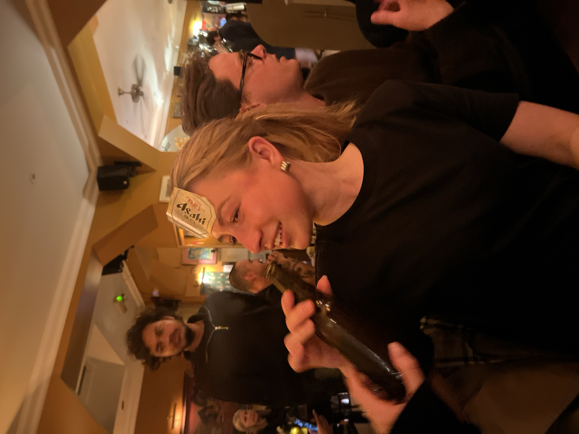 A candid, canted shot shows two women and a man posing in a dimly lit room, likely a bar or restaurant. One of the women has a beer label stuck to her forehead and is holding a dark bottle. All are dressed in dark clothing. The background features a crowd of people, interior decor, and wall-mounted objects suggesting a public gathering place. A ceiling fan is visible in the upper left corner. The location and city are not definitively determined from this single image.