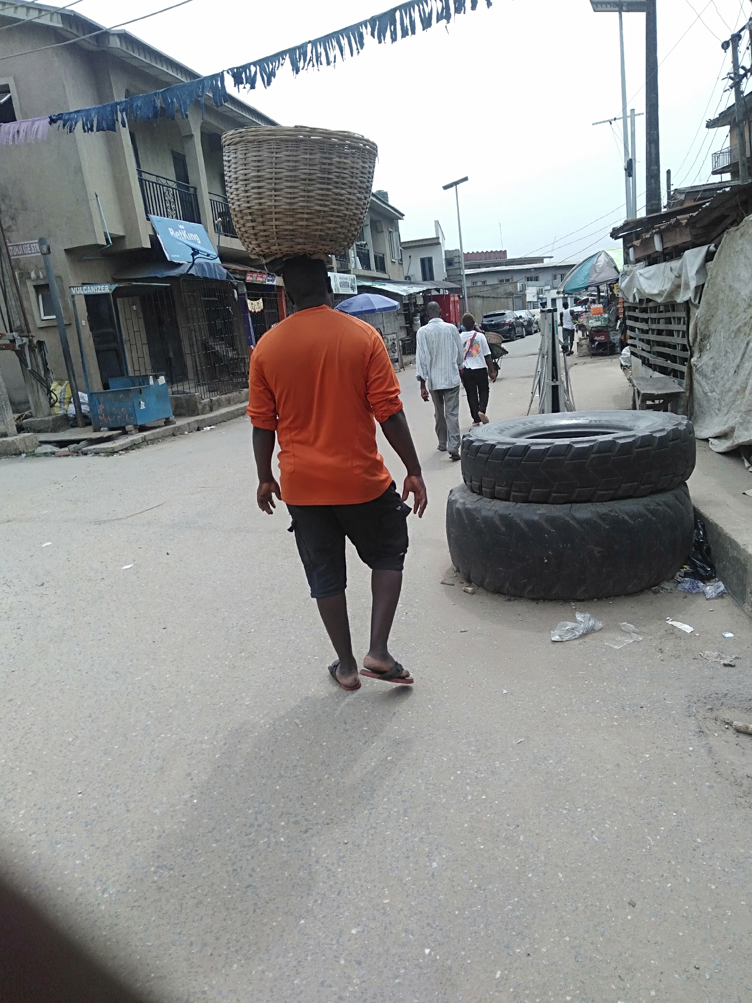 A street scene in Shangisha, Nigeria, shows a man walking away from the viewer, carrying a large woven basket on his head. He is wearing an orange shirt, dark shorts, and sandals. Two other people are walking in the same direction, slightly ahead of him. Stacked against the side of the road are two large tires. The street is paved and there is debris on the ground. Buildings line both sides of the street, with overhead wires visible. Some buildings have awnings. Other figures can be seen further down the street.