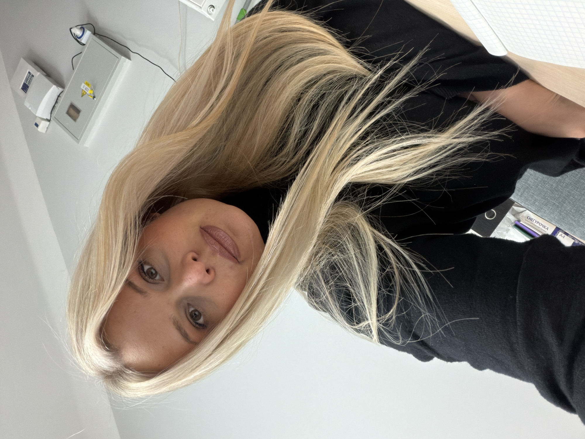 A blonde woman in a black turtleneck sweater is positioned in an indoor environment. She appears to be in a clinical setting, possibly a medical office or clinic, as indicated by the light gray wall and medical equipment visible in the background. The equipment includes what appears to be an electrical panel and a small white device mounted on the wall. Additionally, there's a white surface, possibly a desk or countertop, with paperwork or supplies on it. The woman is looking directly at the camera, suggesting a selfie or a direct portrait taken at close range. The photo shows no specific indications of Sarapul, Russia.