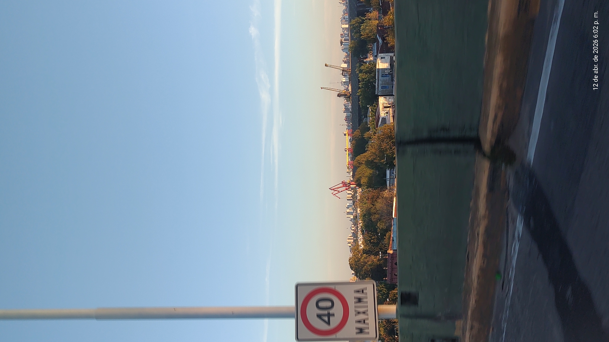 View of Dock Sud, Argentina, from a bridge. A speed limit sign indicating "40 MAXIMA" is in the foreground. Beyond that, a cityscape includes trees, buildings, and cranes. The sky is blue. The bridge's edge and road are visible to the right. Date stamp of "12 de abr. de 2026 6:02 p. m." appears vertically in the upper right.