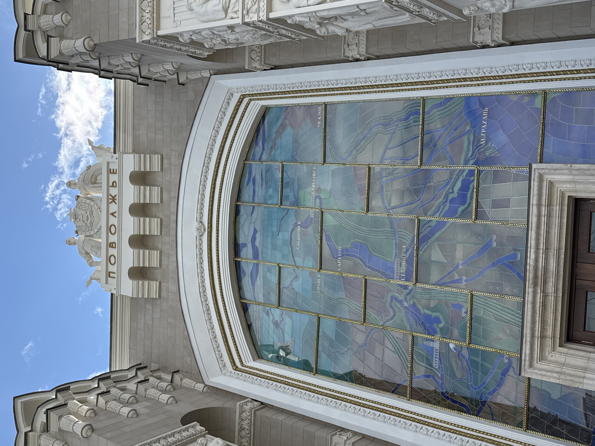 The image shows the facade of the "Volga" pavilion at the All-Russian Exhibition Center (VDNKh) in Moscow, Russia. The pavilion is adorned with classical architectural elements, including columns and sculptures. A large stained-glass window dominates the center of the facade, depicting a map of the Volga River region with various cities labeled in Russian. The inscription "ПОВолЖЬЕ" (Volga Region) is visible above the window. The sky above is blue with scattered clouds. The building's white stone and decorative gold trim create a grand and ornate appearance.