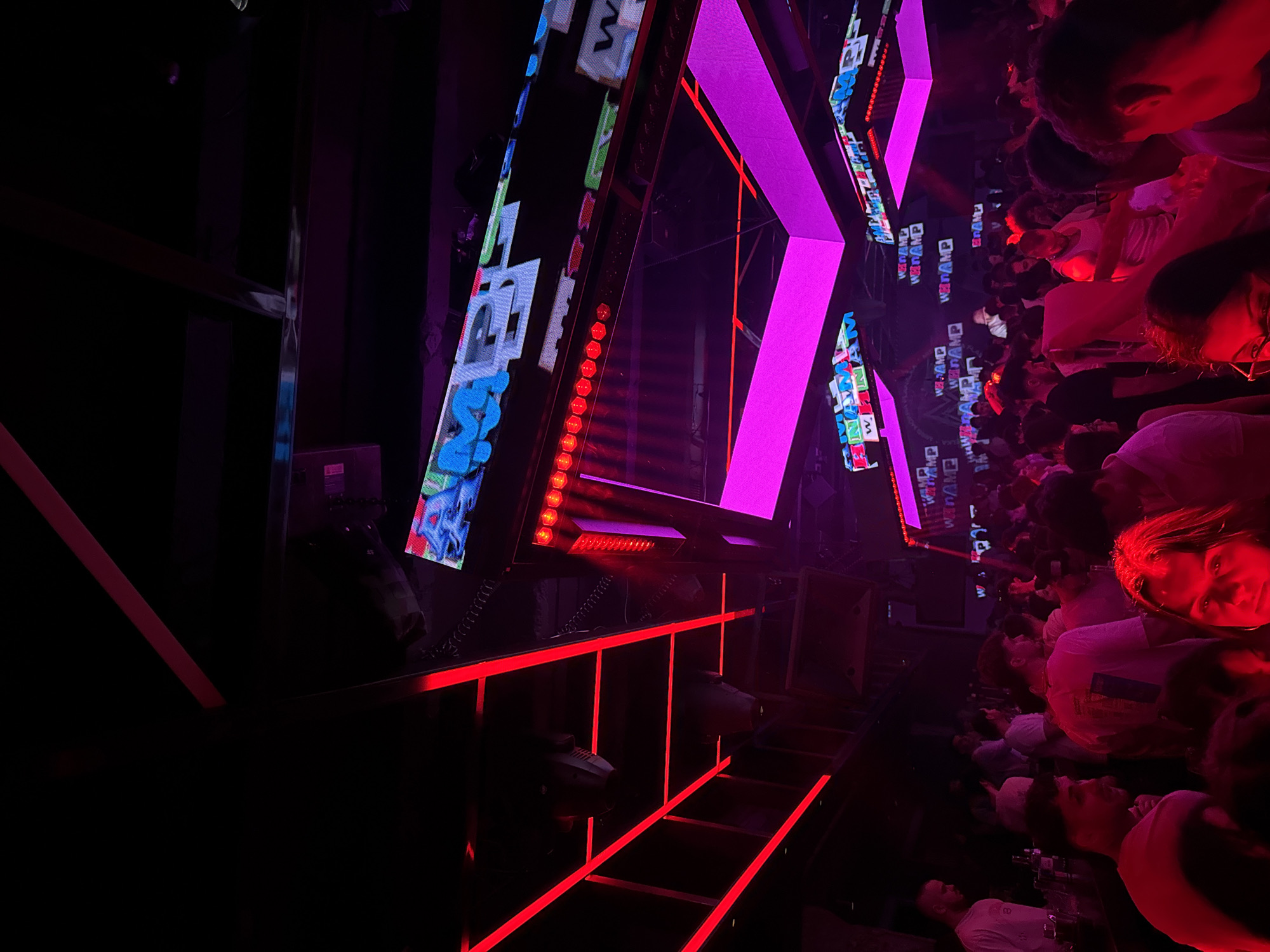 The image depicts a crowded nightclub scene, likely in Sofia, Bulgaria, given the "WEB RAMP" signage visible in Cyrillic script. A large, illuminated structure shaped like an "X" dominates the center of the frame, featuring neon pink outlines and sections of dynamic LED displays with graffiti-like imagery. Red lighting is prominent, particularly in a grid-like structure below the "X" and in the general ambient light. The crowd is dense, with many faces looking upward toward the structure, bathed in the pink and red light. Individuals are casually dressed, suggesting a typical night out. The environment is dark with controlled lighting for effect.