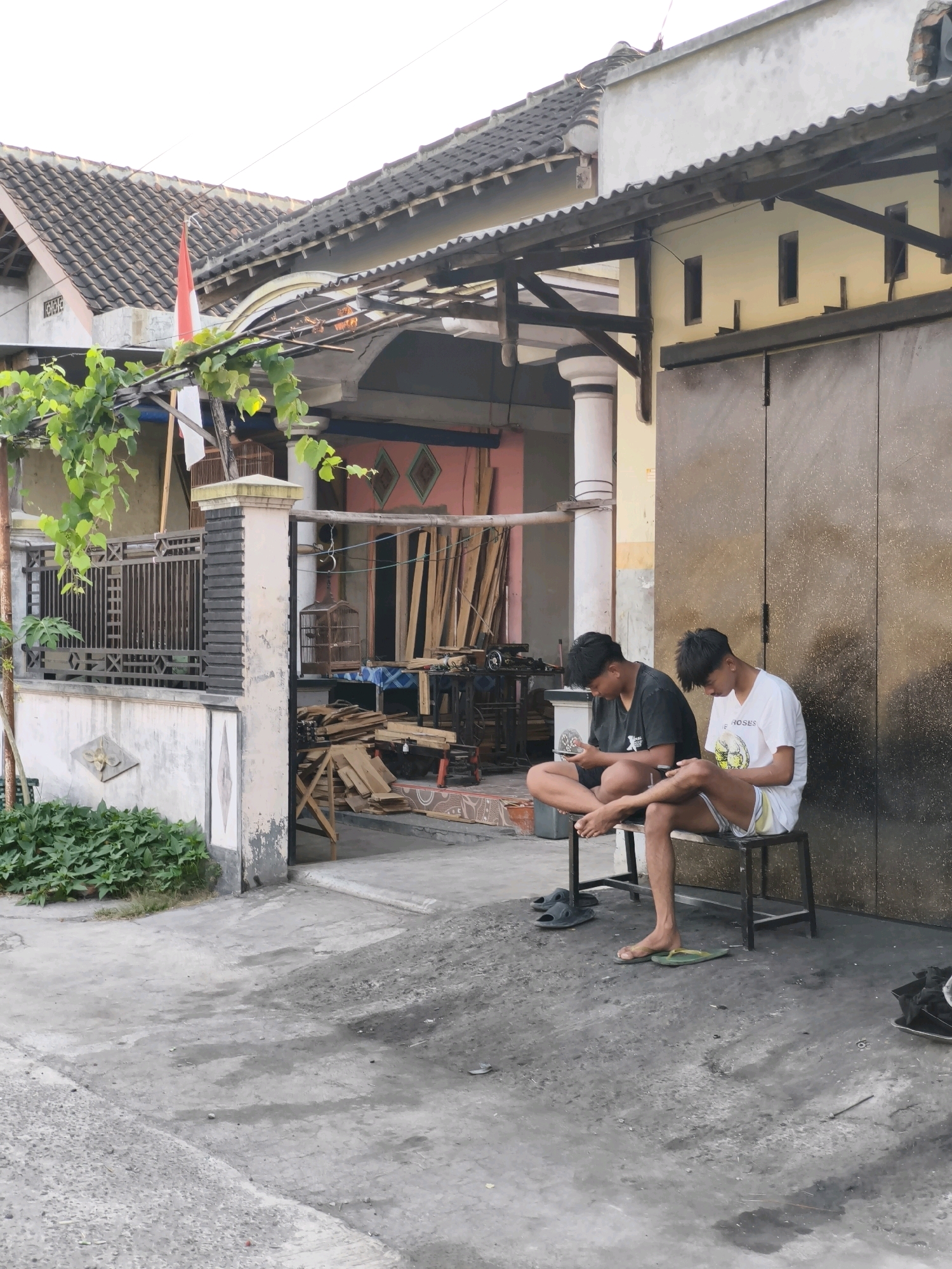 In Desa Dibal, Indonesia, two young males are seated on a low, dark-colored bench in front of a building. Both are engrossed in their mobile phones. The individual on the left wears a black t-shirt and is barefoot, with his legs crossed. The person on the right is in a white t-shirt and shorts, with one foot resting on the ground in a green sandal and the other leg bent. Their footwear, two pairs of dark flip-flops and the green sandal, are on the ground nearby.
The building behind them has a corrugated metal roof and a large, dark, paneled sliding door. The wall to the left of the door is light yellow with rectangular openings. To the left of this structure, a white-painted pillar supports a wooden overhang. A metal fence with decorative patterns encloses an area to the left of the pillar, where a vibrant green plant is growing at the base of a white concrete wall. A small red and white flag, possibly the Indonesian flag, is visible on a pole behind the fence.
The foreground is a concrete paved area with patches of darker, possibly oily, stains and some loose gravel. To the right of the building, a collection of lumber is stacked, and a birdcage hangs. The overall scene suggests an outdoor workspace or residential area.