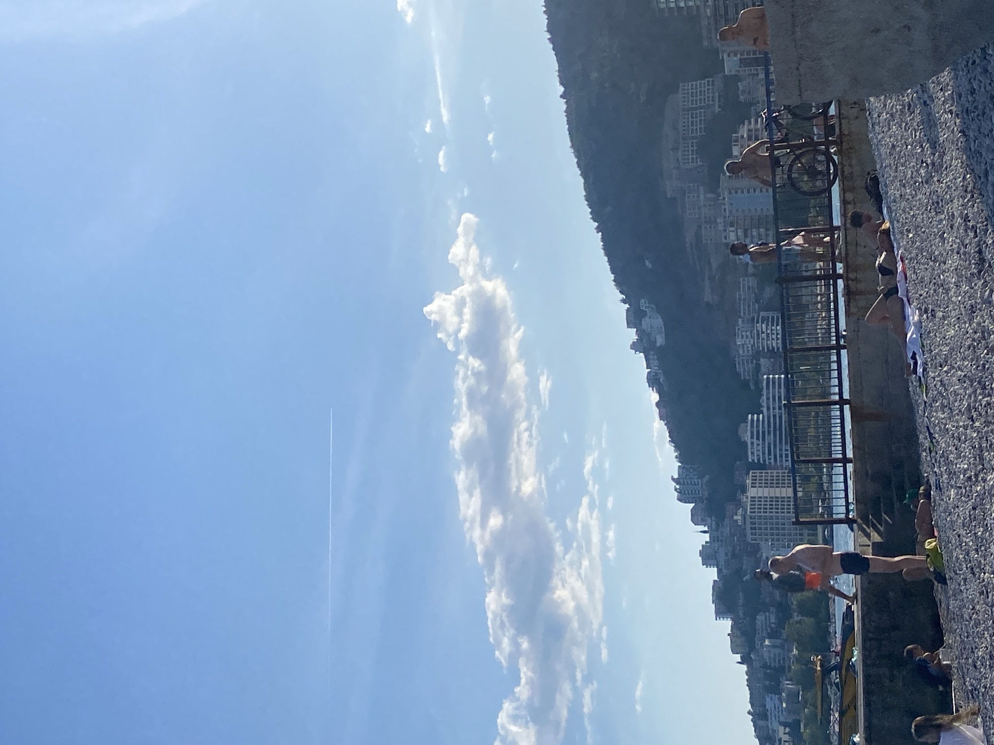 The image captures a sunny day in Budva, Montenegro, possibly at a seaside location near the city center. A group of people are seen enjoying the day by the water. A man in dark shorts is diving or preparing to dive off a stone ledge that is lined with a metal railing. Other people are standing near the railing, while some are lying down, seemingly sunbathing. A bicycle is parked on the ledge next to the railing. Behind the ledge, buildings are visible on a hillside. The sky is mostly clear blue with a contrail and a long, vertical cloud formation in the center. The overall environment suggests a recreational scene in an urban setting along the coast.