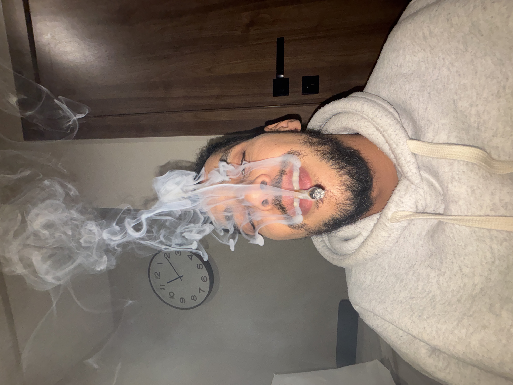 A male individual is depicted indoors, framed from the upper chest upwards, with a lit cigarette or joint held in his open mouth. Dense white smoke billows upwards from his mouth, partially obscuring his face, dark beard, and mustache. His eyes appear closed or squinted due to the smoke. He is wearing a light grey hooded sweatshirt with a visible drawstring. In the background, to the left of the subject, a light-colored wall features a round analog wall clock showing the time as approximately 12:15. To the right of the subject, a dark wooden door with a black handle and a square black lock plate is visible. The specific location within the city of Tanger, Morocco, cannot be determined from the visual evidence presented in the image.