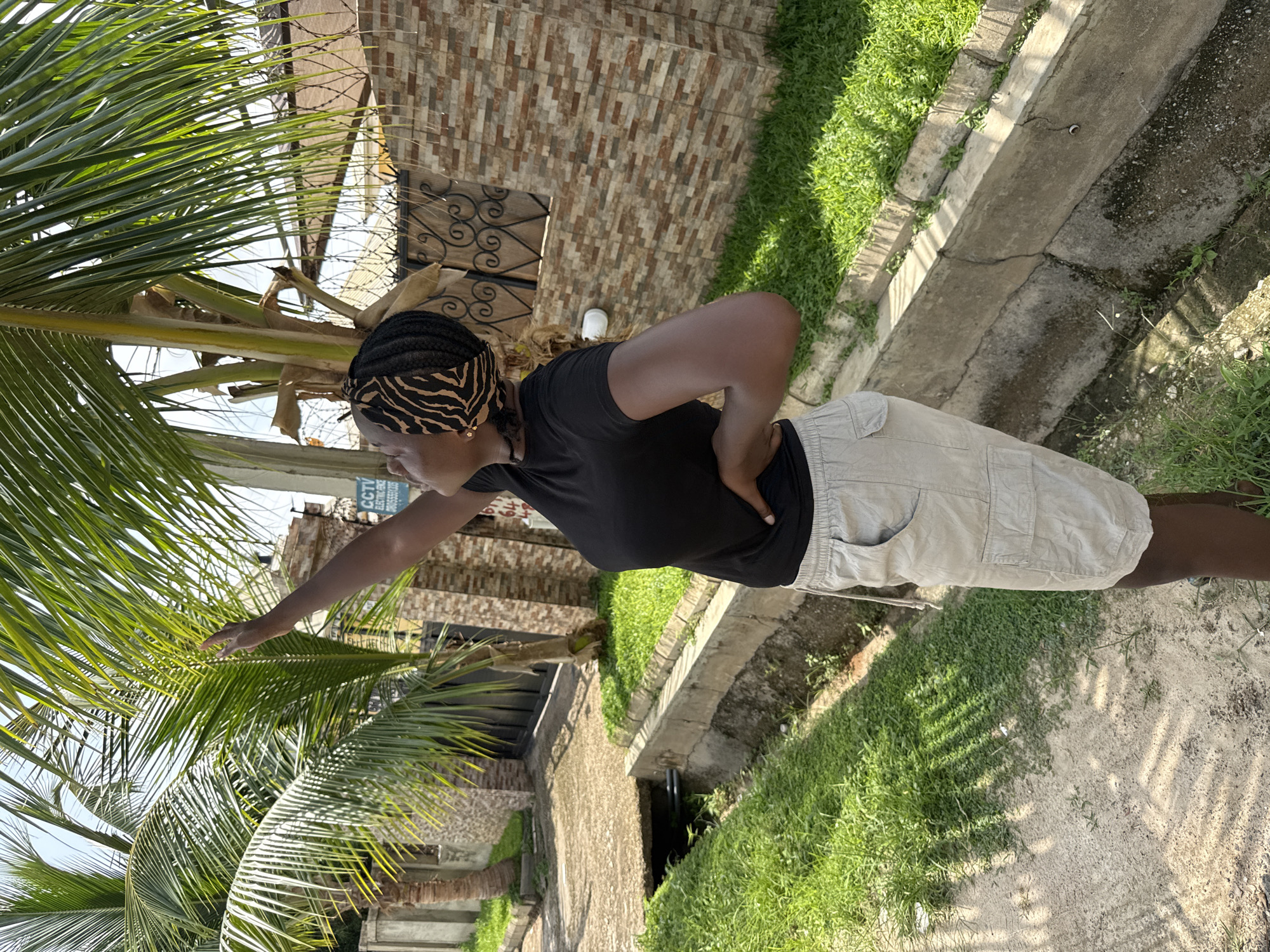 A woman stands on a walkway near a palm tree, possibly in Abuja, Nigeria. The woman is wearing a black shirt, khaki shorts, and a patterned head wrap. Her left hand is outstretched toward the palm tree. A building with a decorative iron gate is visible behind her. A narrow concrete drainage ditch runs parallel to the walkway. Grass grows on either side of the ditch. The ground is a mixture of concrete and dirt. The sky is visible through the palm fronds.