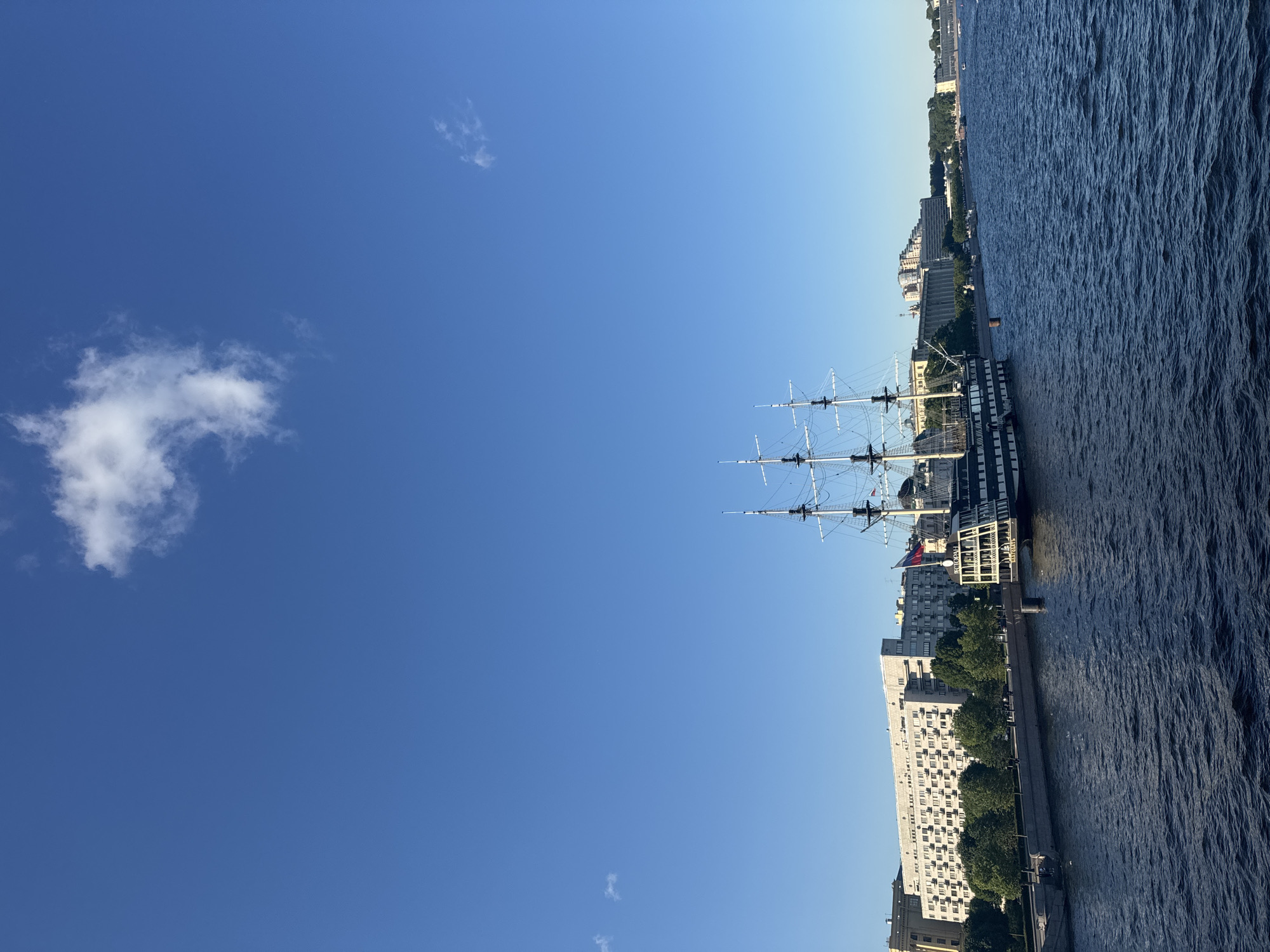 The image shows a tall ship docked on a river in front of a city skyline. The sky is a clear blue with a few wispy white clouds. The ship is a traditional sailing vessel with three masts and rigging. It is dark in color with white trim and appears to be a museum ship or a historical replica.
Behind the ship, a waterfront promenade runs along the river. Beyond the promenade, there are several multi-story buildings, typical of urban architecture. Trees line the waterfront, providing a touch of greenery. In the distance, more buildings and trees can be seen along the river, suggesting a sprawling city. The river itself is wide and appears to be choppy, with visible ripples on the surface. The overall scene is one of a historic ship in an urban setting, under a bright, clear sky.