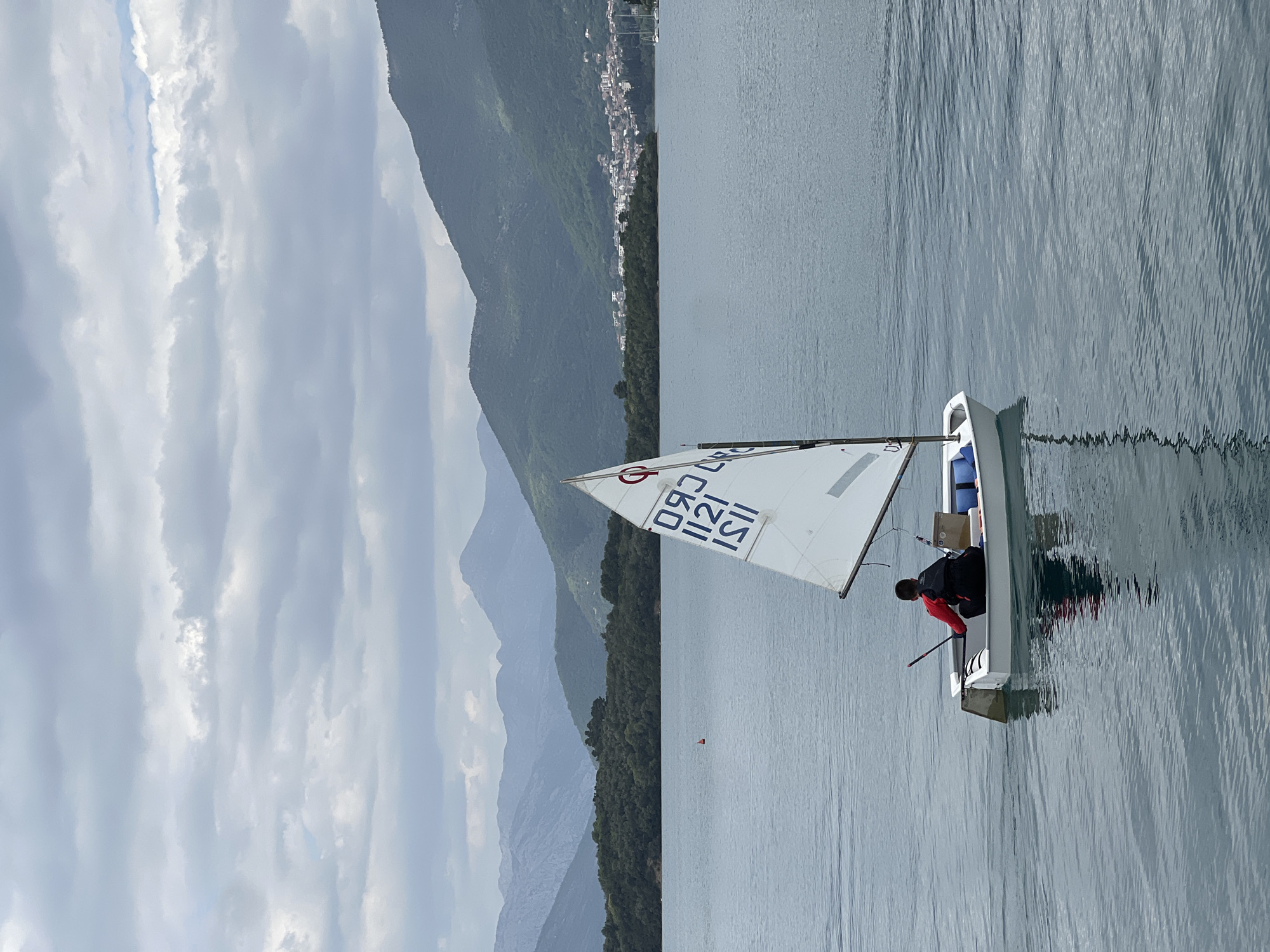 The image depicts a tranquil sailing scene on a body of water in Đuraševići, Montenegro, under an overcast sky. A single person, dressed in a red and black jacket, is seated in a white dinghy sailboat in the foreground. The sail, predominantly white, displays blue and red markings, including the letters "ISL" (likely a country code) and the numbers "1121" stacked below them. Another number, "0832" (or "Z830" in reverse), is visible higher on the sail.
The water is calm, showing slight ripples and reflections of the sky and the boat. A small red buoy is visible floating in the water to the left of the sailboat in the mid-ground. In the background, majestic, forested mountains dominate the landscape, receding into hazy layers. Along the base of the nearest mountain range, a cluster of white buildings indicates a small town or village. The sky above is filled with soft, light grey clouds, suggesting an overcast day. The overall impression is one of peaceful watersports activity in a scenic natural setting.