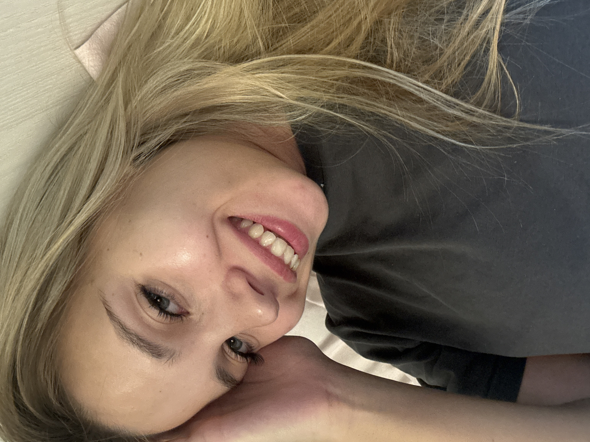 A young woman with blonde hair and blue eyes smiles into the camera, her left hand resting on her cheek. She is wearing a dark grey t-shirt. The background appears to be a wall with a textured pattern and a pink fabric object. The lighting suggests an indoor setting, possibly a bedroom. There is no discernible indication of the location within Chaikovsky, Russia.