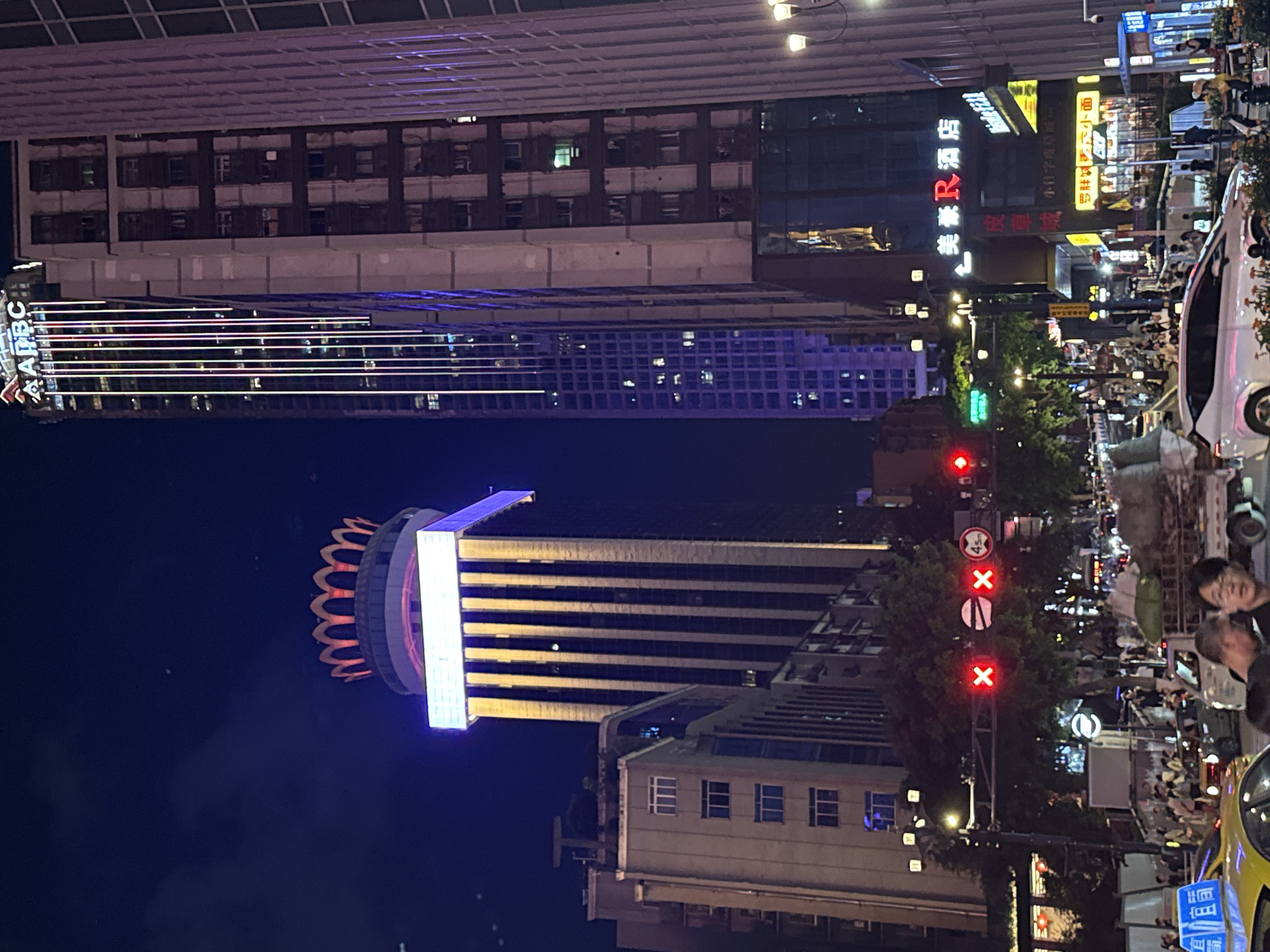 This image captures a nighttime street scene in Yuzhong District, China, dominated by towering illuminated buildings. The architecture is modern, with skyscrapers featuring intricate lighting designs. One prominent building is topped with a crown-like structure emitting a warm orange glow, while another displays vertical lines of colorful LED lights. The street below is bustling with traffic, including cars and a truck loaded with goods. Pedestrians are visible on the sidewalks, and various illuminated signs indicate shops and businesses. Traffic lights, displaying red signals, are also present. The overall atmosphere is one of a vibrant urban night.