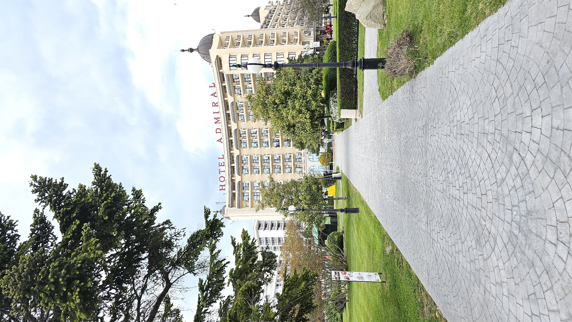 The image is a ground-level, outdoor shot of the Hotel Admiral in Varna, Bulgaria, taken on a sunny day. The hotel is a tall building with a beige facade, topped with a dome and ornate features. The name "HOTEL ADMIRAL" is vertically displayed in red letters. A paved walkway leads towards the hotel, flanked by green lawns, trees, and trimmed hedges. Black lampposts line the walkway. A stone sculpture is visible on the lawn, as well as a few people in the distance. A food advertisement board can be seen on the right side of the frame. The sky is blue with some white clouds.