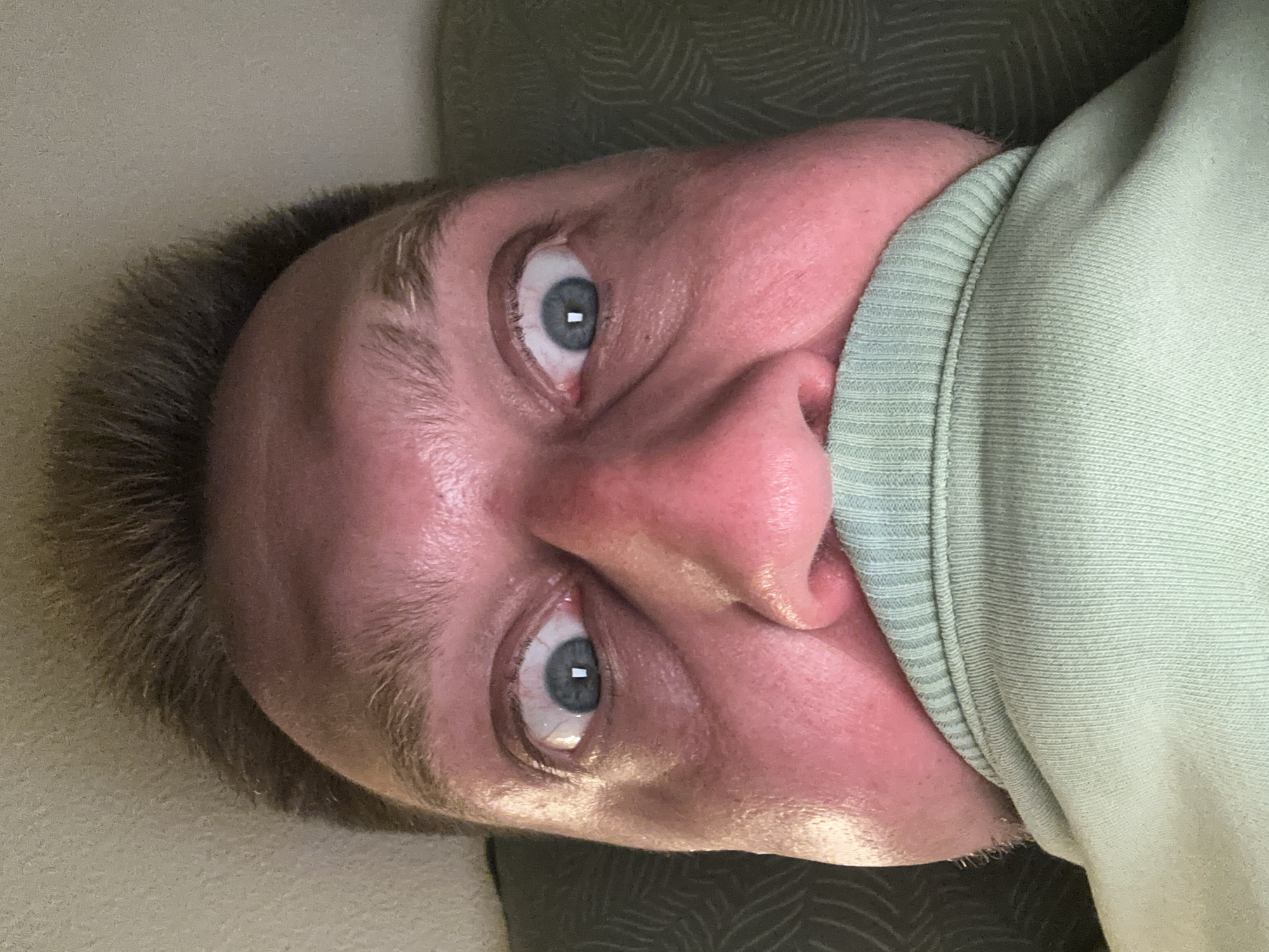 The image is a close-up, high-angle, selfie-style photograph of a person's face, with their lower face obscured by a light green garment, possibly a sweatshirt. The person's eyes are wide open and appear to be looking directly at the camera. The skin is flushed, and there is visible sweat on the forehead and cheeks, suggesting a warm environment or physical exertion. The background consists of a textured surface, likely a wall, and a patterned fabric, possibly a couch or bed. The lighting appears to be artificial and possibly from a flash, as indicated by the glare on the eyes. There is no information in the image to suggest a location in Zvezdara Urban Municipality, Serbia.