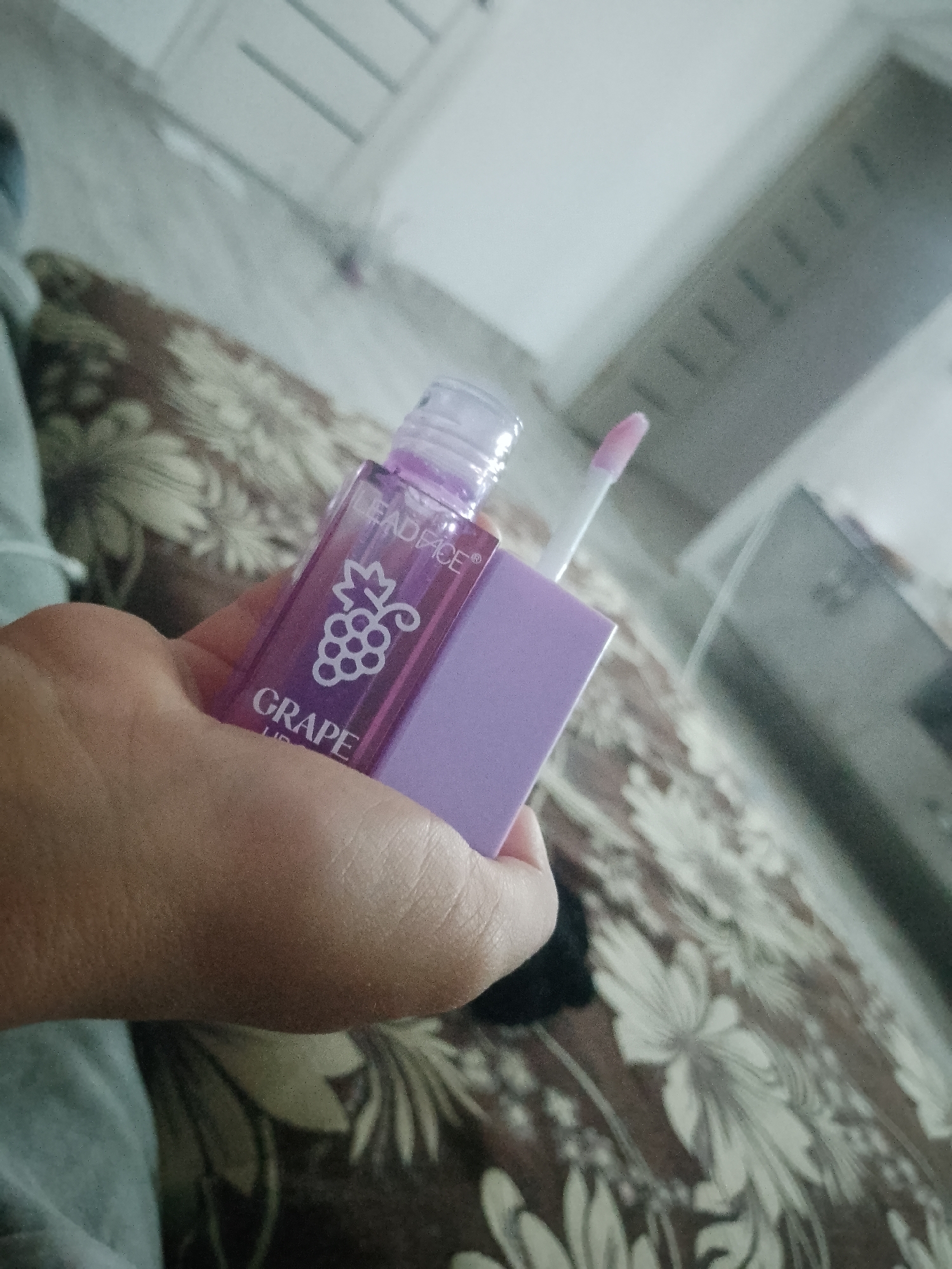 A person's hand is holding a clear purple lip gloss bottle with a purple cap. The label on the bottle is white and features a drawing of a bunch of grapes and the text "LEAD FACE" and "GRAPE". The lip gloss wand is extended from the bottle. The background is a patterned fabric surface and a blurred reflection of what appears to be a room with furniture. The image is taken indoors and is well-lit. The location in the city of Келдемұрат, Kazakhstan is not discernible from the image.