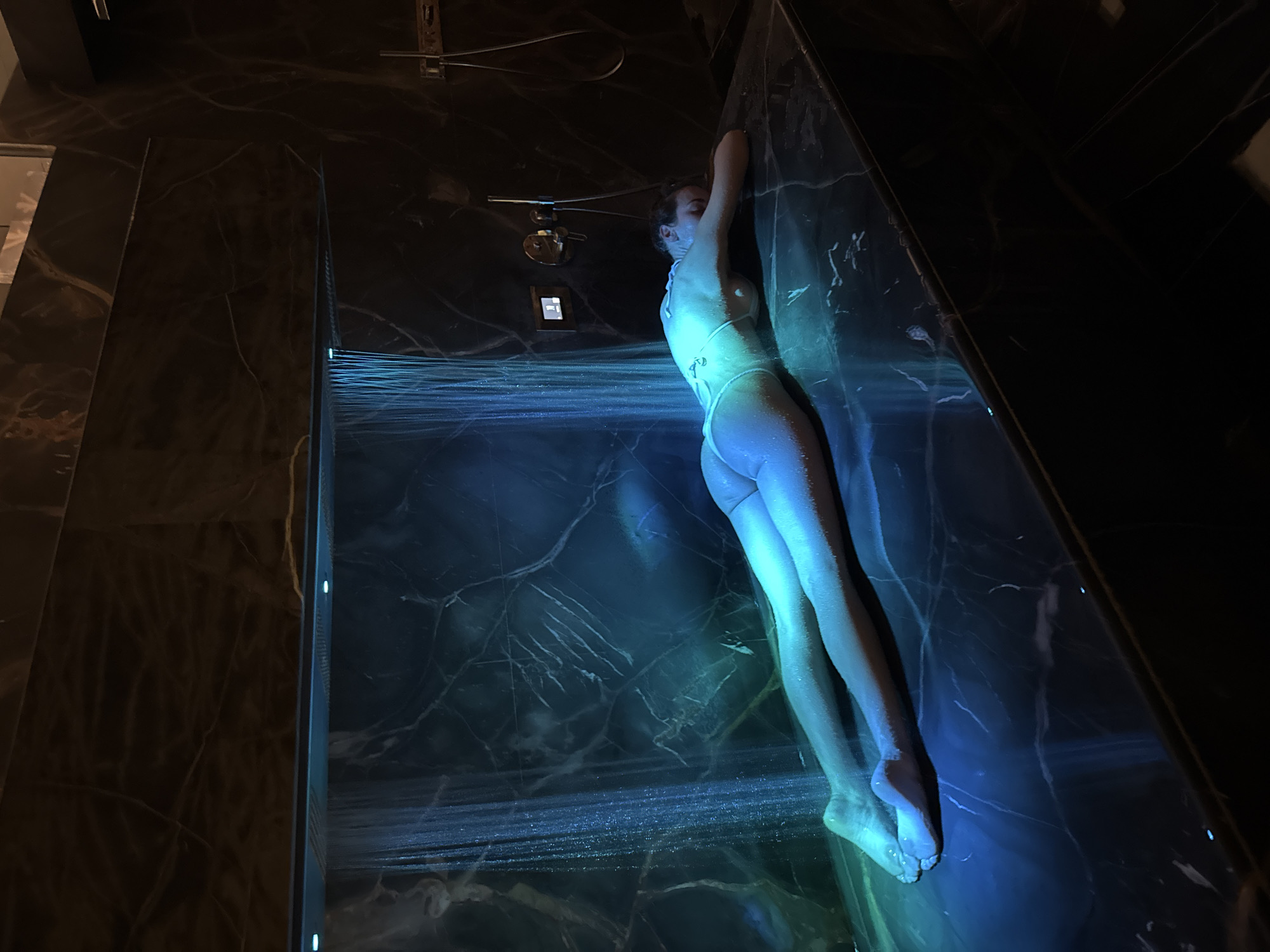 This image captures a single adult female lying prone on a dark, polished marble slab within what appears to be a luxurious private spa or shower area. The specific location is Unknown, Unknown.
The woman is positioned horizontally across the slab, face down, with her head turned slightly to the right and her arms extended forward, hands resting near her head. She is wearing a light-colored two-piece swimwear, and her body is wet, glistening under the artificial illumination.
Above her, a long, linear shower fixture is recessed into the dark marble wall, emitting multiple vertical streams of water, creating a waterfall effect. This shower fixture incorporates blue LED lights, which cast a prominent blue and green hue onto the falling water, the woman's wet body, and the surrounding marble surfaces.
The walls and the surface the woman rests on are composed of dark, highly polished marble, exhibiting distinct, light-colored veining. To the right of the shower stream, on the marble wall, chrome-finished shower controls are visible, including a circular mixing valve, a square digital display or control panel, and a mounted handheld shower hose and head. The overall environment is dimly lit, with the blue-green light from the shower serving as the primary source of illumination, creating a serene and atmospheric scene.
