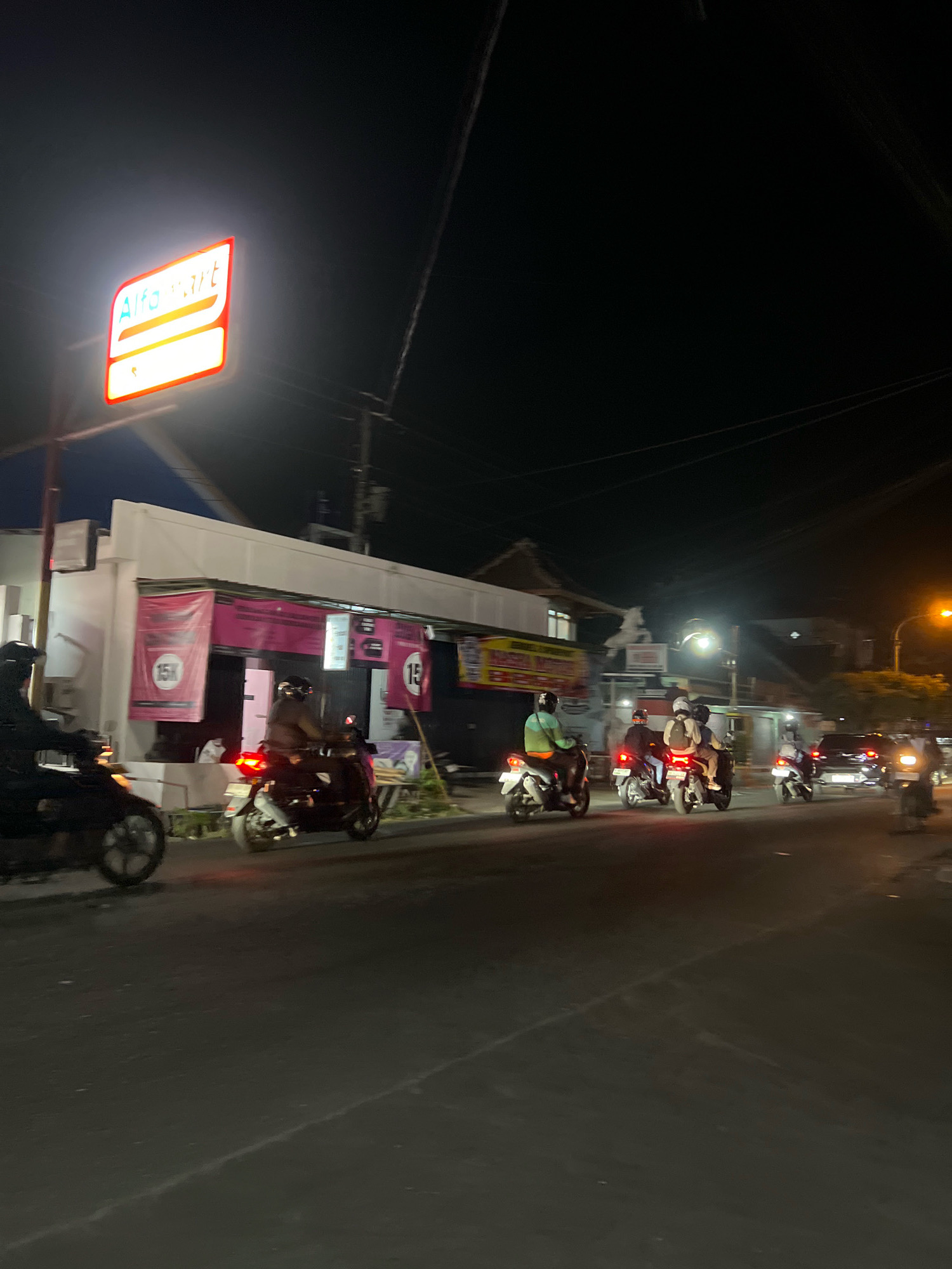 This image depicts a nighttime street scene in Tlogoadi, Indonesia, likely in front of an Alfamart convenience store. Several individuals on motorcycles are present, with some riding and others parked. The Alfamart store is identifiable by its illuminated red and white sign. Pink banners with text and "15K" markings are visible on the store's facade. The street is paved and appears to be a public thoroughfare with streetlights and electrical wires overhead. The overall atmosphere suggests a busy urban environment at night.