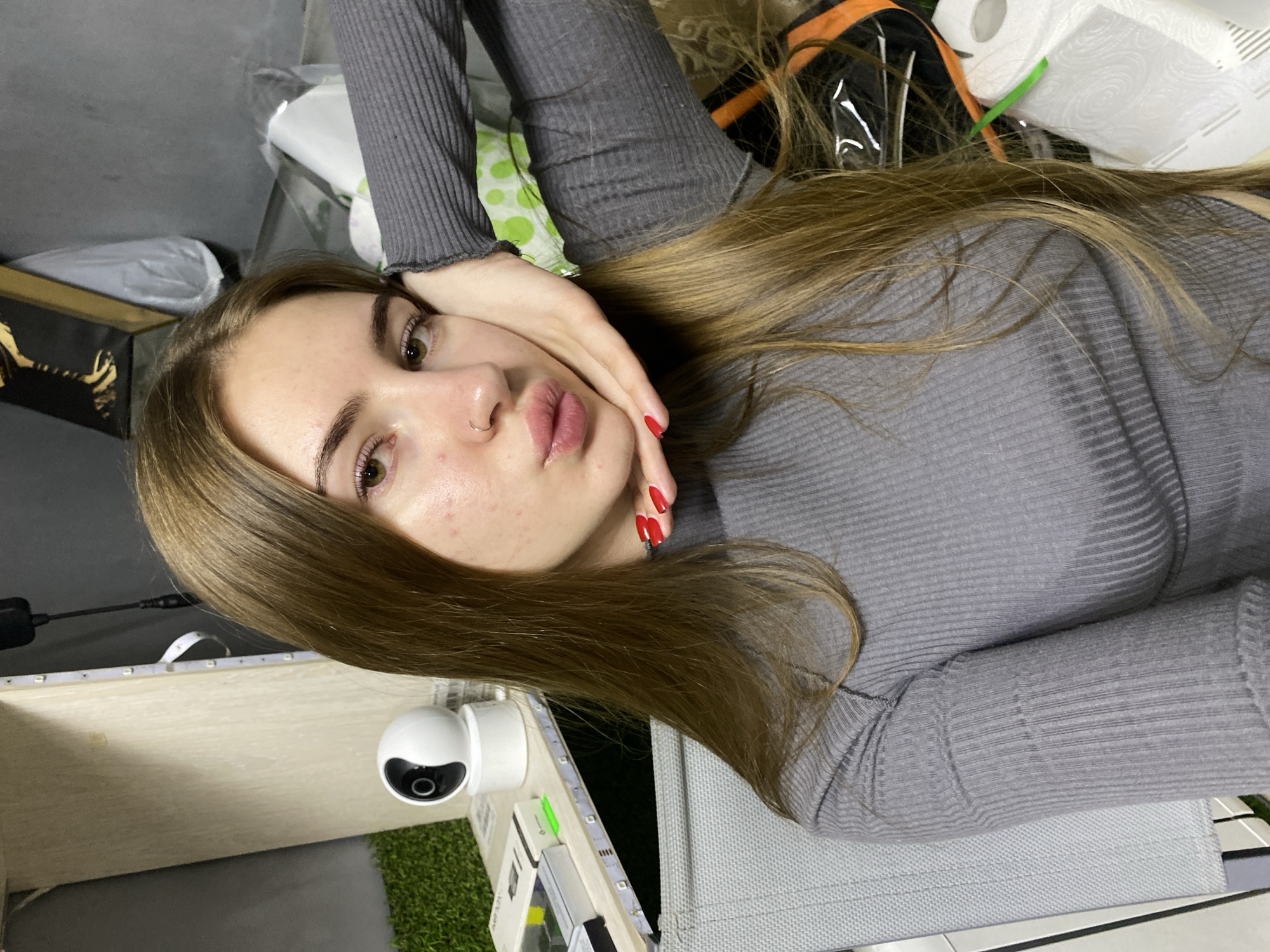 A young Caucasian woman with long, straight, light brown hair and brown eyes is taking a selfie indoors. She is wearing a grey, ribbed, long-sleeved shirt. Her right hand is resting on her cheek, and her lips are pursed. Her fingernails are painted bright red. She has a subtle nose ring.
The background appears to be a cluttered indoor space. To her left, a white spherical security camera sits on a light-colored shelf unit that has LED strip lighting attached. A green, artificial grass mat is visible behind the camera. In the upper right, a framed artwork with a golden abstract design is hanging on a gray wall. Various objects, including white plastic packaging, and rolls of paper towels are visible. The overall impression is of a casual, informal setting. The specific location within the city of Дьяково, Russia, cannot be determined from the visual information provided.