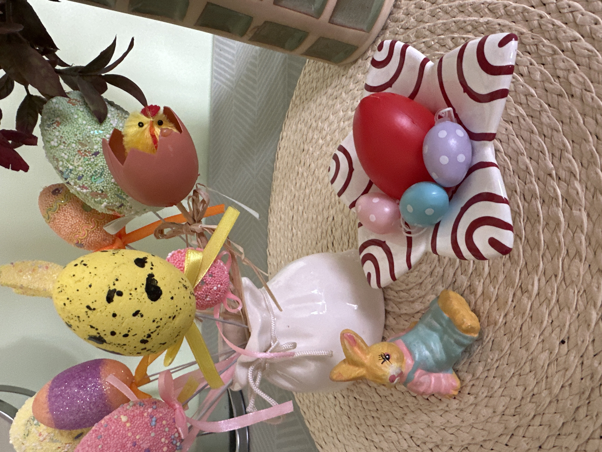 The image depicts an Easter-themed decorative display. Several artificial Easter eggs in various colors and textures are arranged with ribbon. A small, yellow, chick-in-egg decoration is present. A ceramic ornament featuring red and white swirls is adjacent to colorful miniature eggs. A ceramic bunny figurine is present. A woven mat or hat serves as a background. The location is not evident.