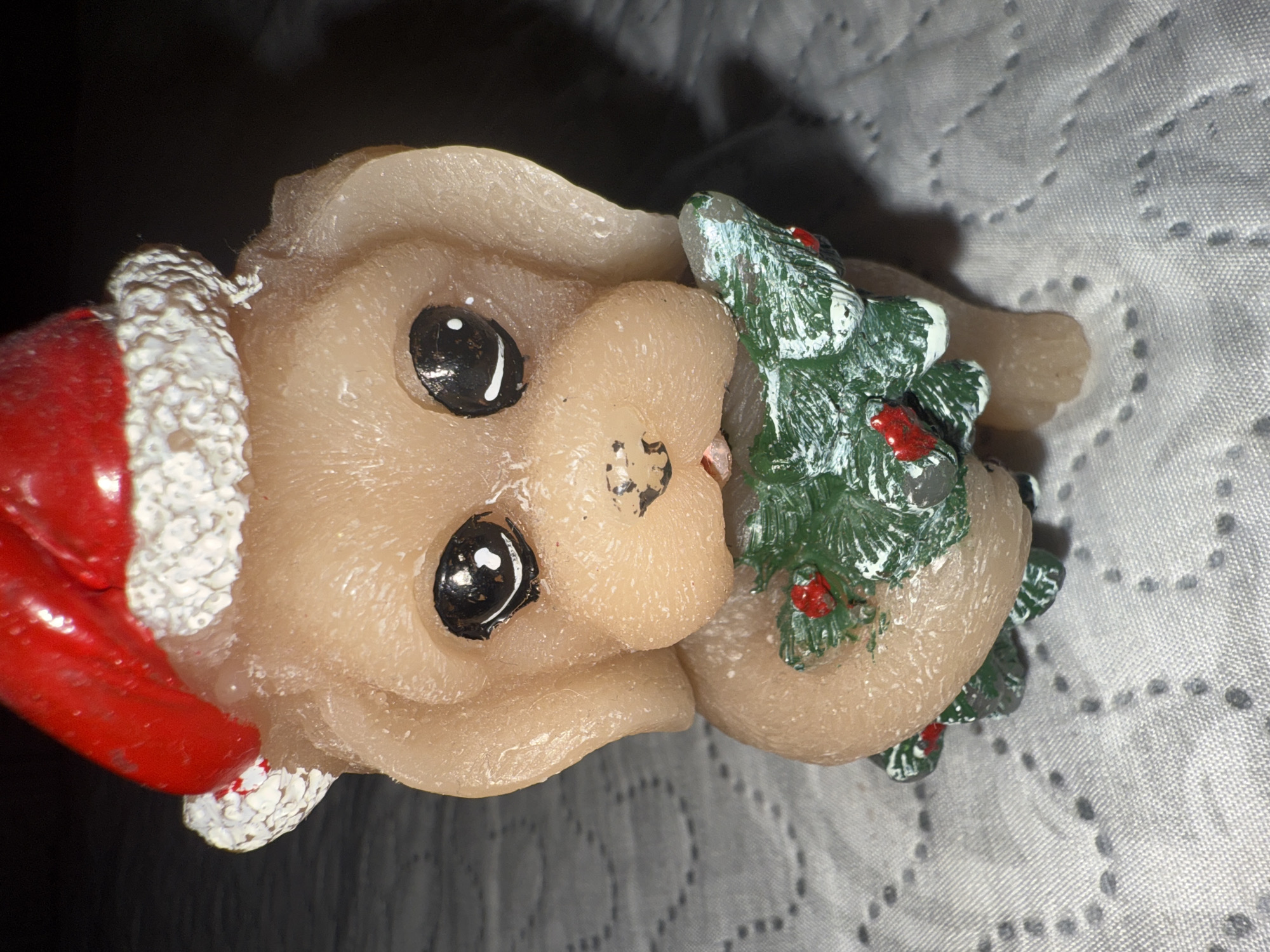 The image presents a close-up view of a festive Christmas figurine, likely a decorative item or candle, positioned diagonally on a light-colored, patterned fabric surface. The central subject is an animal-like figure, possibly a piglet or rabbit, with a smooth, light beige or cream-colored body. It is adorned with a vibrant red Santa hat, featuring a textured white trim and a white pompom at its tip. The figurine's black eyes possess distinct white highlights, imparting a cheerful and whimsical expression. A small, slightly reddish indentation marks the nose area.
The figure is depicted holding a miniature green Christmas tree, which is decorated with white "snow" accents and small red berries or ornaments. The material of both the figurine and the tree appears slightly waxy or resinous, with visible textures and some minor imperfections, suggesting a handcrafted quality.
The background consists of a light-colored, possibly white or light grey, fabric with a subtle, repeating pattern of dark, circular dots or indentations, resembling a quilted surface. Shadows are cast behind the figurine, indicating an indoor setting with a distinct light source. The overall scene conveys a holiday theme, characterized by a cute and festive aesthetic. No text is visible in the image. The location context of Varna, Bulgaria is provided, but no visual cues in the image indicate this specific geographical location.