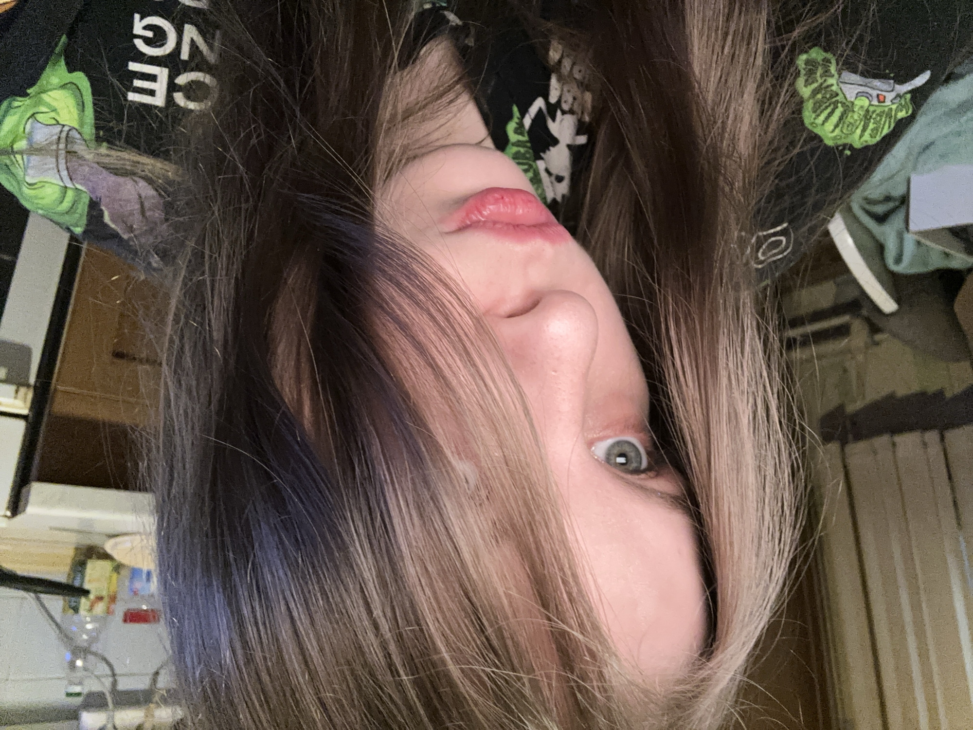 A close-up, upside-down image shows a person with long, light-brown hair that is parted in the middle. Darker roots are visible. Only one eye, which is a gray-green color, is fully visible. The person's nose and lips are visible as well, the lips are pink. The person is wearing a black shirt with white and green graphics. Behind the person, there is a partially visible room with a desk, some items on it and light in the background. There is no evidence to indicate a location in Moscow, Russia.