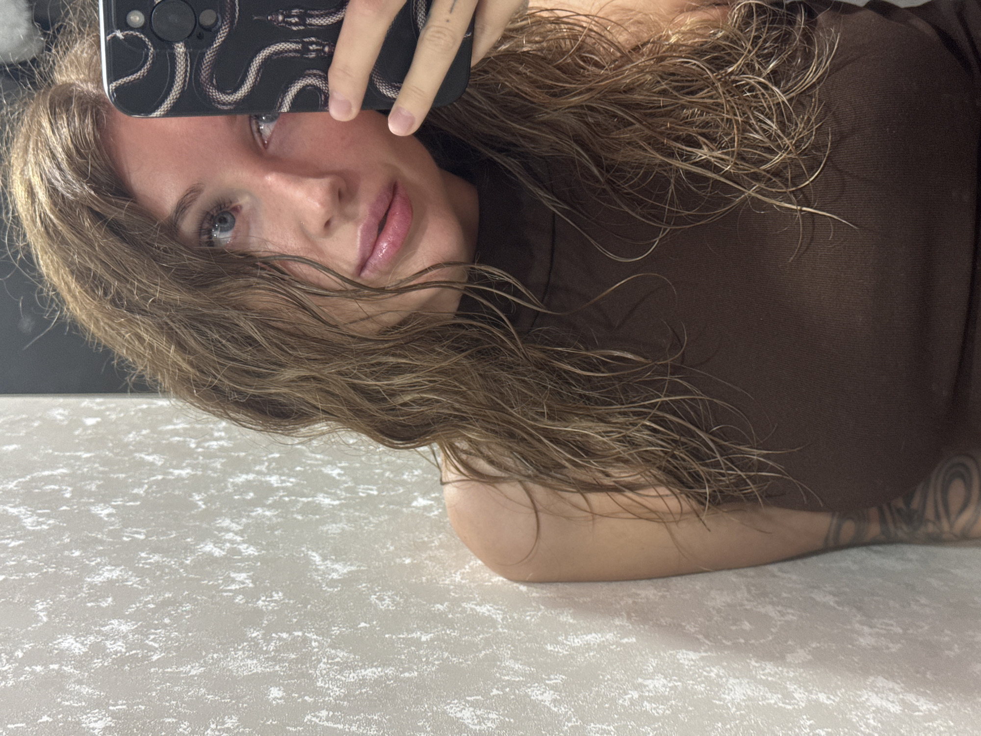 The image shows a person taking a selfie with a phone. The person has long, brown hair and is wearing a brown top. They have blue eyes and pink lips. A tattoo is visible on their arm. The background is a light-colored surface with a speckled pattern. The lighting suggests it is daytime. The phone case has a black and white snake design. The image is cropped closely to the subject, with their face and upper body visible. The overall tone is casual and personal. There is no visible text in the image.