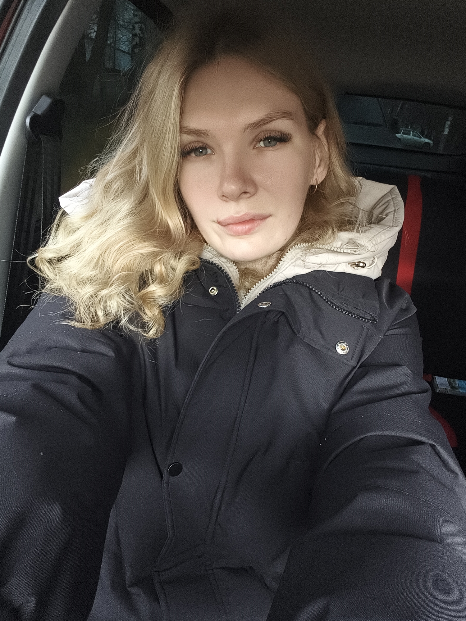 A blonde woman is sitting in the driver's seat of a car. She is wearing a dark blue winter coat with a beige hood. The interior of the car is dark. The reflection in the rearview mirror appears to show a light-colored car. The location cannot be determined from the image.