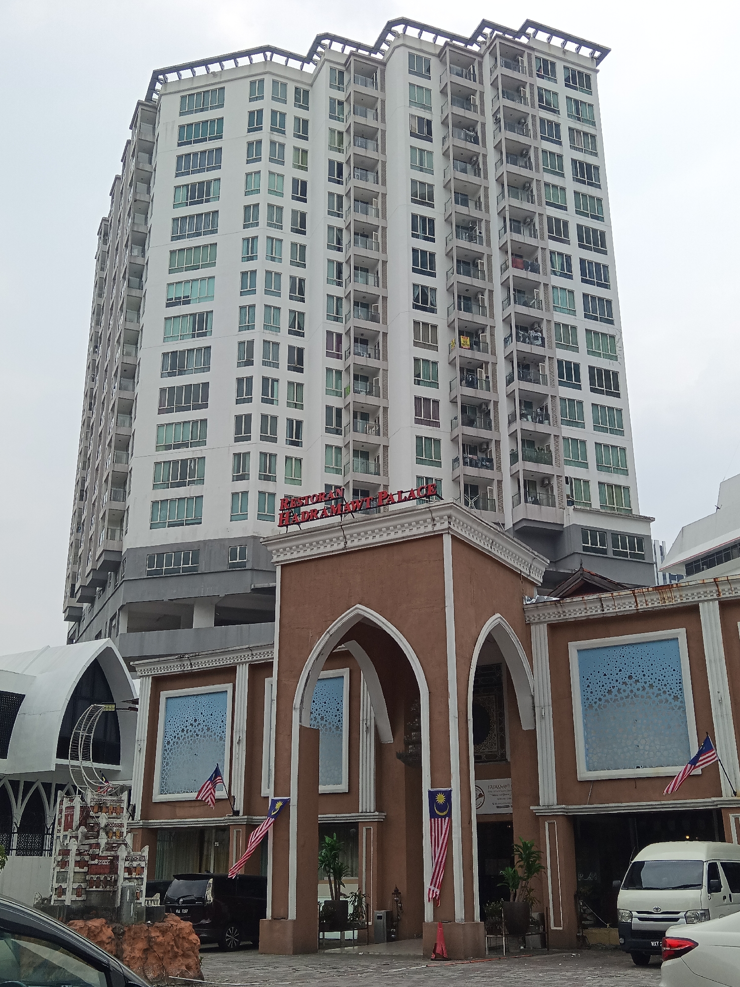 The image shows a multi-story residential or commercial building in Kuala Lumpur, Malaysia. The building is tall and white, with many windows and balconies. In the foreground, there is a building with a brown facade and arched entrances. Two Malaysian flags are visible, one hanging from the brown building and another on a flagpole. There is also a white van and a black car parked in front of the building. The sky is overcast. The sign above the arched entrance reads "RESTORAN HAILAM CHU PALACE". The building appears to be located in a commercial area, possibly with shops or restaurants on the ground floor. The overall scene suggests a typical urban environment in Malaysia.