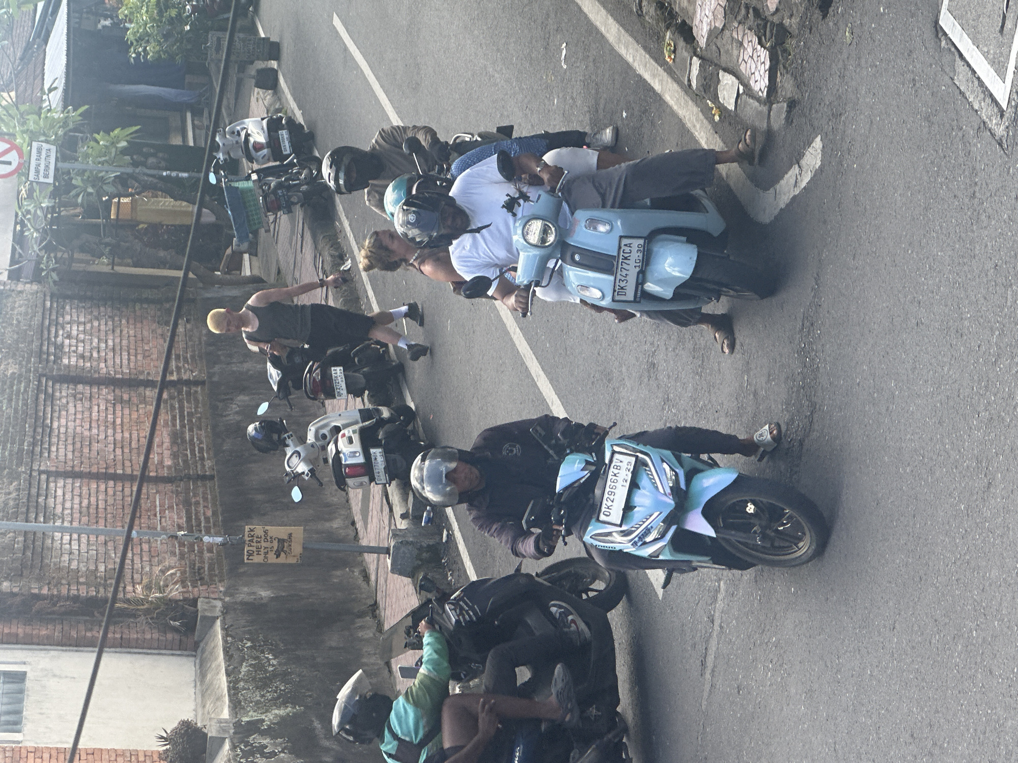 This is an eye-level, medium shot of a street scene in Ubud District, Indonesia. The image captures several people riding on scooters, navigating a paved road with a white line marking a lane. On the right side of the road, there is a sidewalk with patterned paving stones and some small offerings of flowers. Buildings with brick and concrete construction are visible in the background, along with some vegetation and street signs.
In the foreground, a person in a dark jacket and helmet rides a light blue scooter with the license plate "DK2956KBV". To their left, another person is riding a dark scooter, wearing a teal jacket and a helmet.
Further down the street, a person in a white t-shirt and dark pants rides a light blue scooter with the license plate "DK3477KCA". They are accompanied by two passengers, one wearing a helmet and the other with blonde hair. Behind them, a person wearing a beige shirt and helmet rides a scooter.
On the sidewalk to the left, a man with blonde hair wearing a dark tank top and shorts is walking. Behind him, several scooters are parked or in motion. One scooter, white with a black seat, has the license plate "DK8725KAR". Another scooter, white with a blue seat, has the license plate "DK6770WBK".
A sign on a pole reads "NO PARK HERE ONLY DOG IS OK SUKMA". Another sign with a red circle and a diagonal line indicates traffic rules. The overall atmosphere suggests a busy street with typical daily activity in a tropical urban environment.