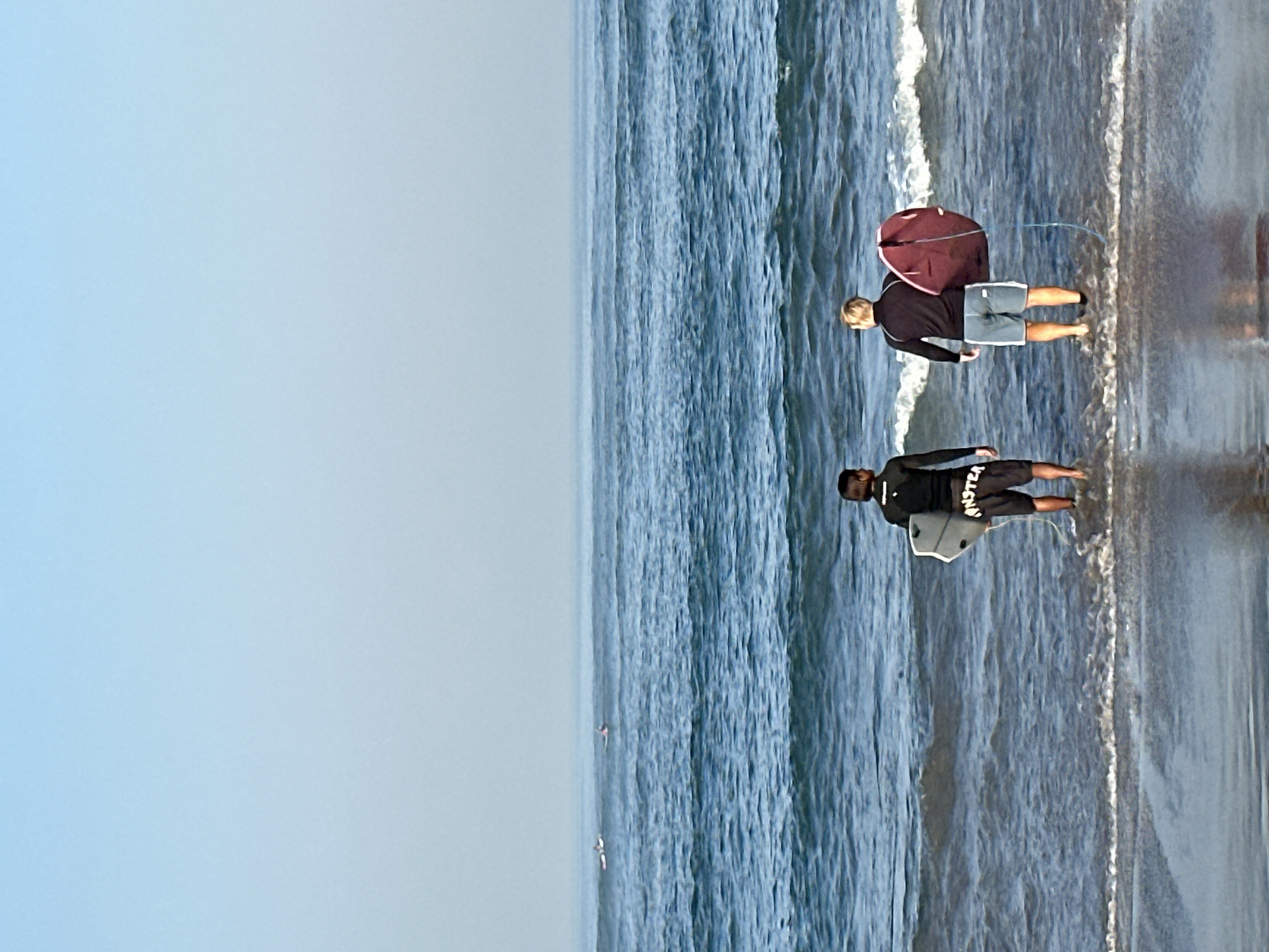Two individuals, viewed from behind, stand at the edge of the ocean. The person on the left wears a black long-sleeved rash guard and black shorts with white lettering, holding a white surfboard. The person on the right wears a black long-sleeved shirt and light blue shorts, carrying a dark red, oval-shaped board with a blue leash. Both are partially submerged in shallow water, facing the sea. The ocean stretches to the horizon, with small waves breaking near the shore. In the distance, several other surfers are visible on their boards. The sky is a clear, pale blue, transitioning to a lighter shade near the horizon. The beach in the foreground is wet, reflecting the sky and figures. The scene appears to be located at a beach in North Kuta, Indonesia.