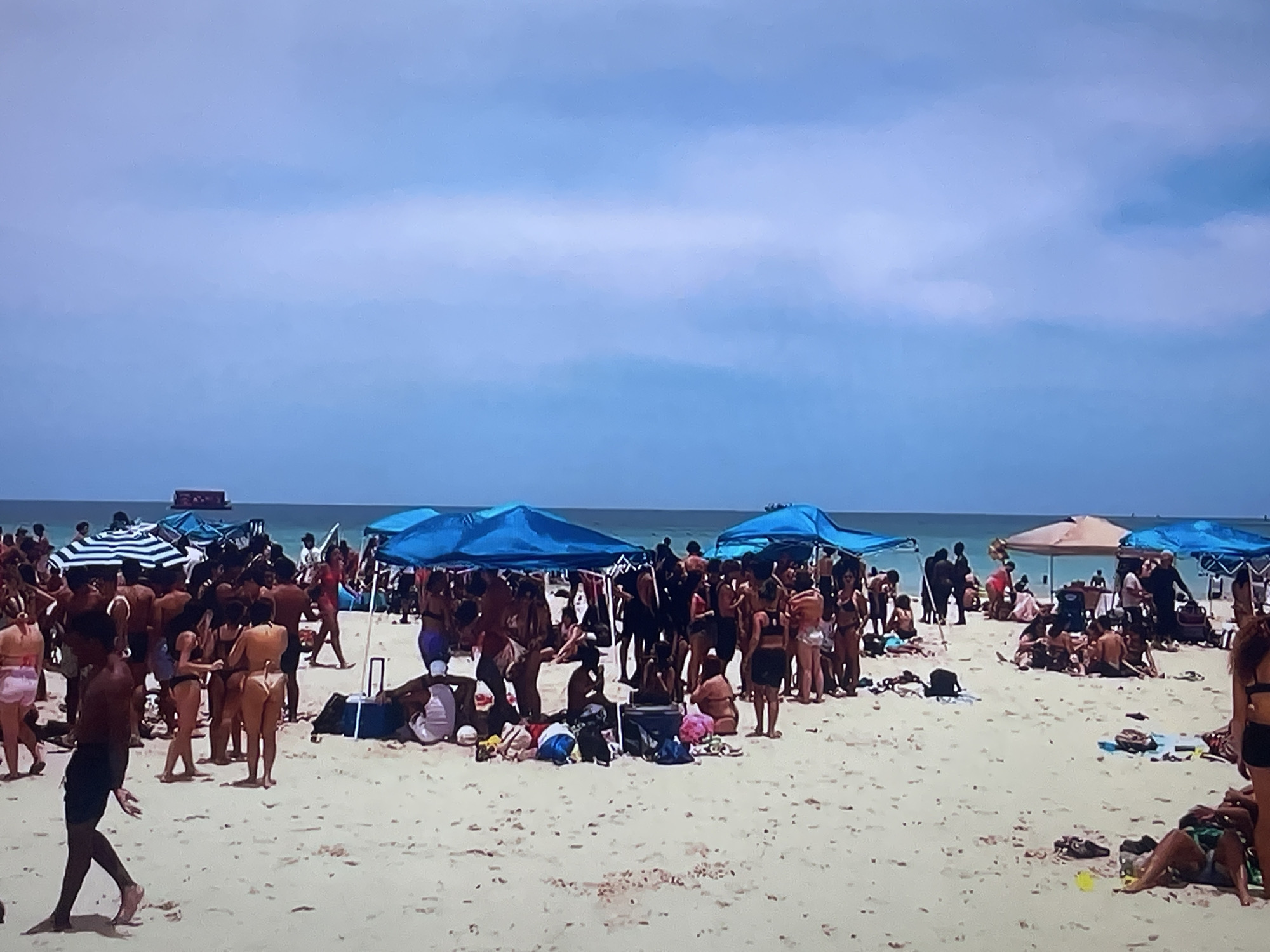 The image depicts a crowded beach scene under a clear blue sky with scattered clouds. Numerous people are scattered across the sandy beach, many in swimwear. Several beach umbrellas and tents provide shade. In the background, the ocean stretches to the horizon. The image does not provide any information that would indicate the location is Shakhovskaya, Russia.
