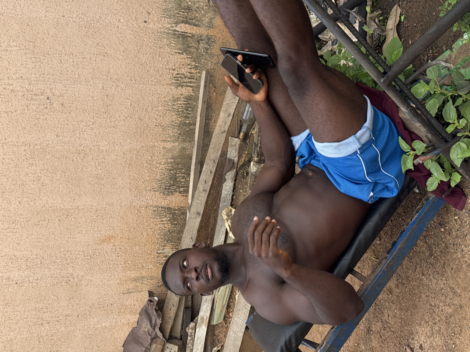 In Akwanga, Nigeria, a Black man is lying on a weight bench outdoors. He is shirtless and wearing blue athletic shorts. His left arm is resting on his stomach, and his right arm is extended, holding two cell phones. The man is looking upwards and to his left. He is positioned in front of a textured, peach-colored wall. To the left of the man, there are stacks of wooden planks and some debris. The ground is dirt and gravel, with some green plants growing near the bench. The weight bench is made of dark metal and has a black padded seat.