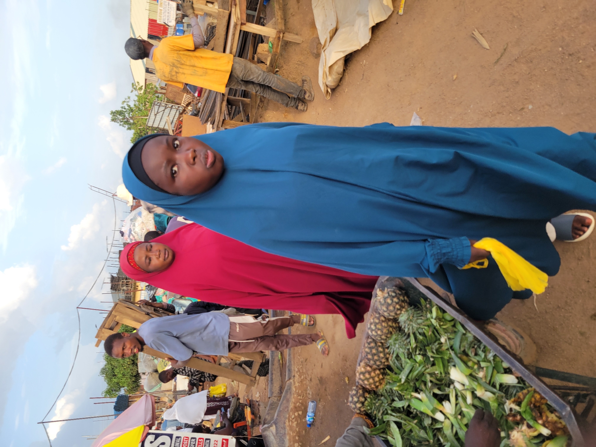 The image depicts a street scene in Gura Top, Nigeria. In the foreground, a young person wearing a blue hijab and dress is looking directly at the camera. To their left, a woman in a red hijab and dress is also visible. A man in a light blue shirt and brown pants is walking away from the camera carrying a wooden structure. In the background, other people, market stalls, and buildings can be seen under a cloudy sky. A cart filled with pineapples and green leaves is also present in the foreground. The ground is sandy and dusty, typical of a market environment.